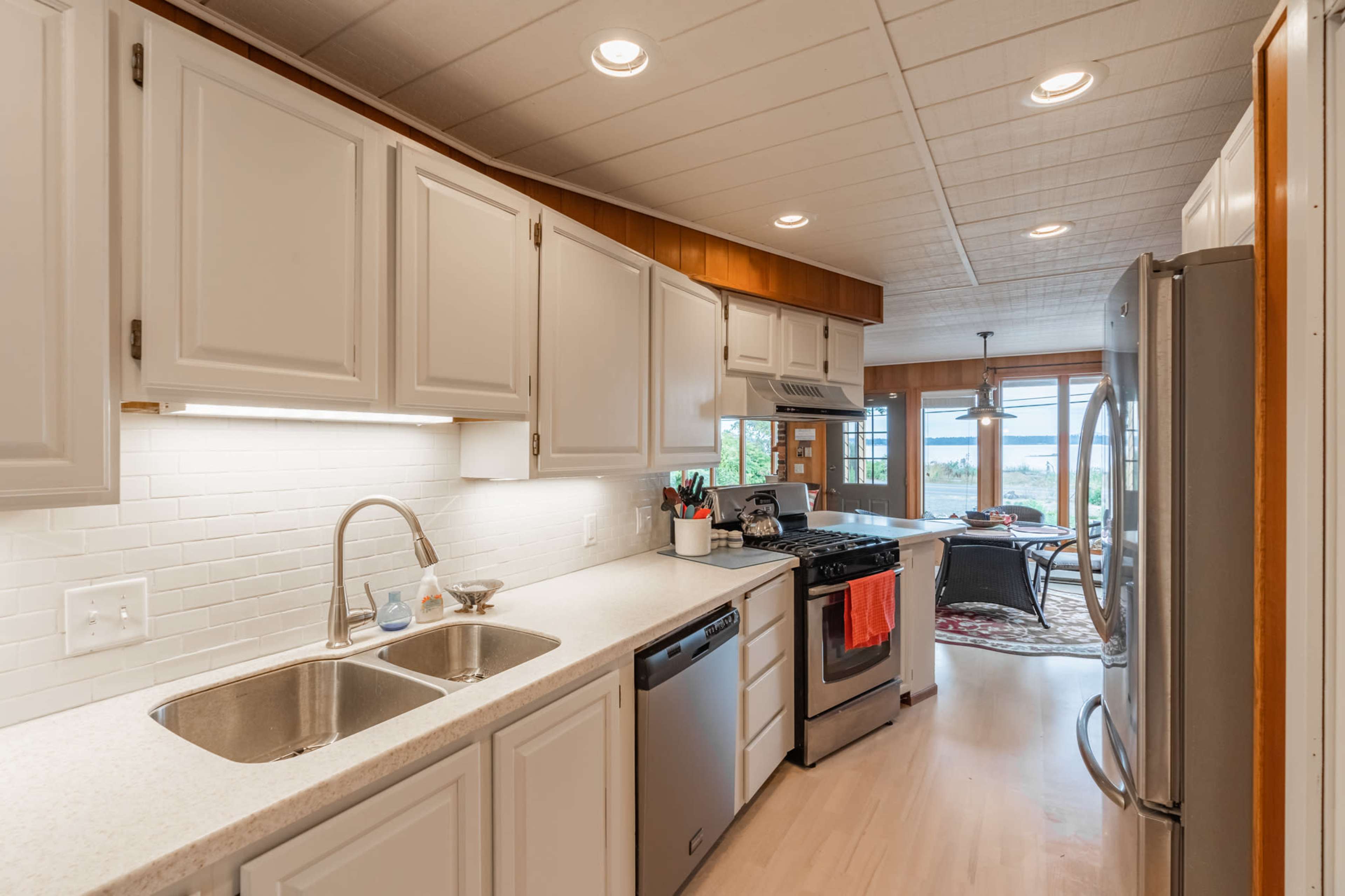 The image shows a modern kitchen with white cabinets, a double sink, and stainless steel appliances, leading to a dining area with large windows.
