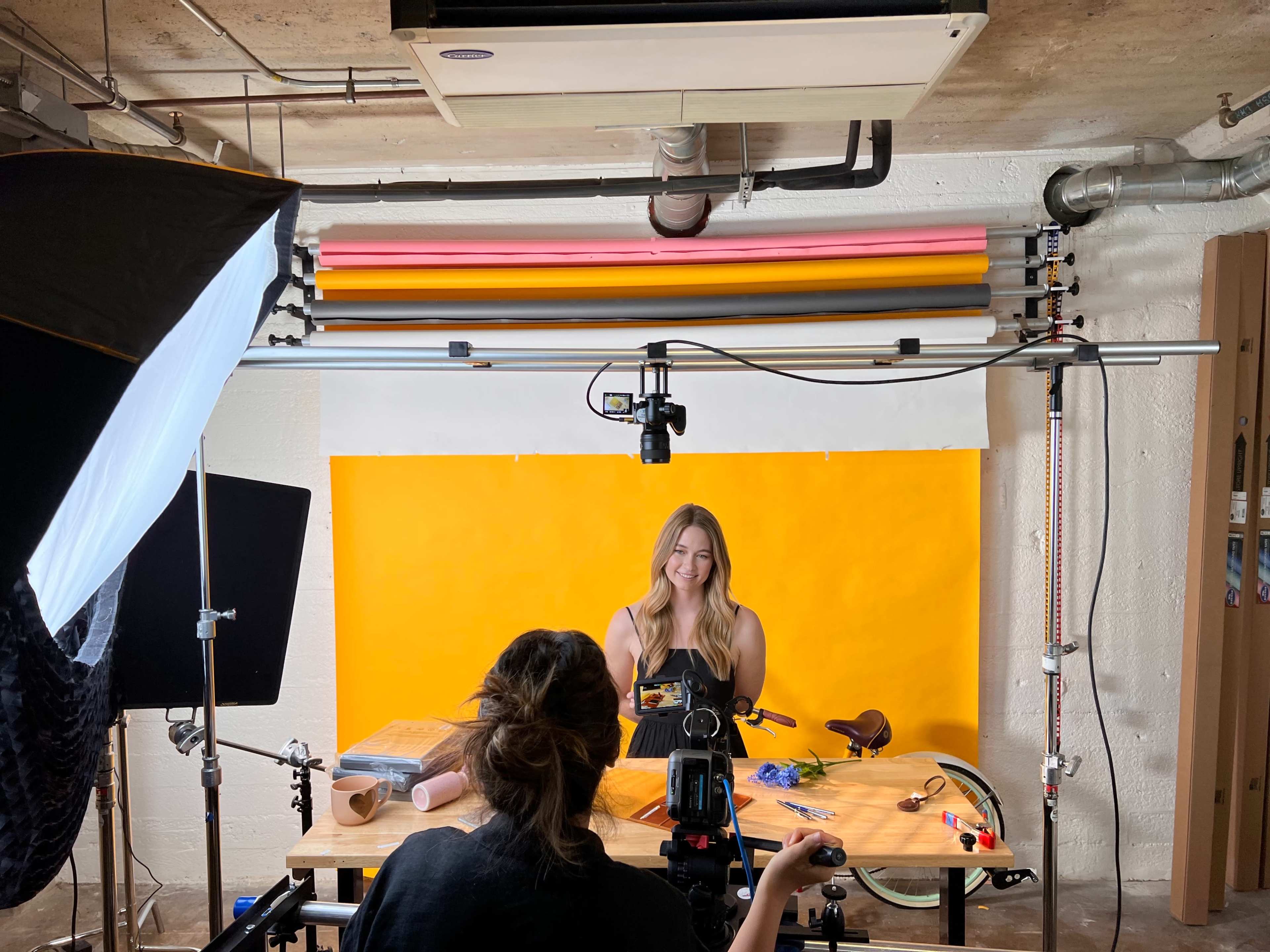A woman is being filmed in a studio setting with a bright yellow backdrop, while another person operates a camera on a tripod.