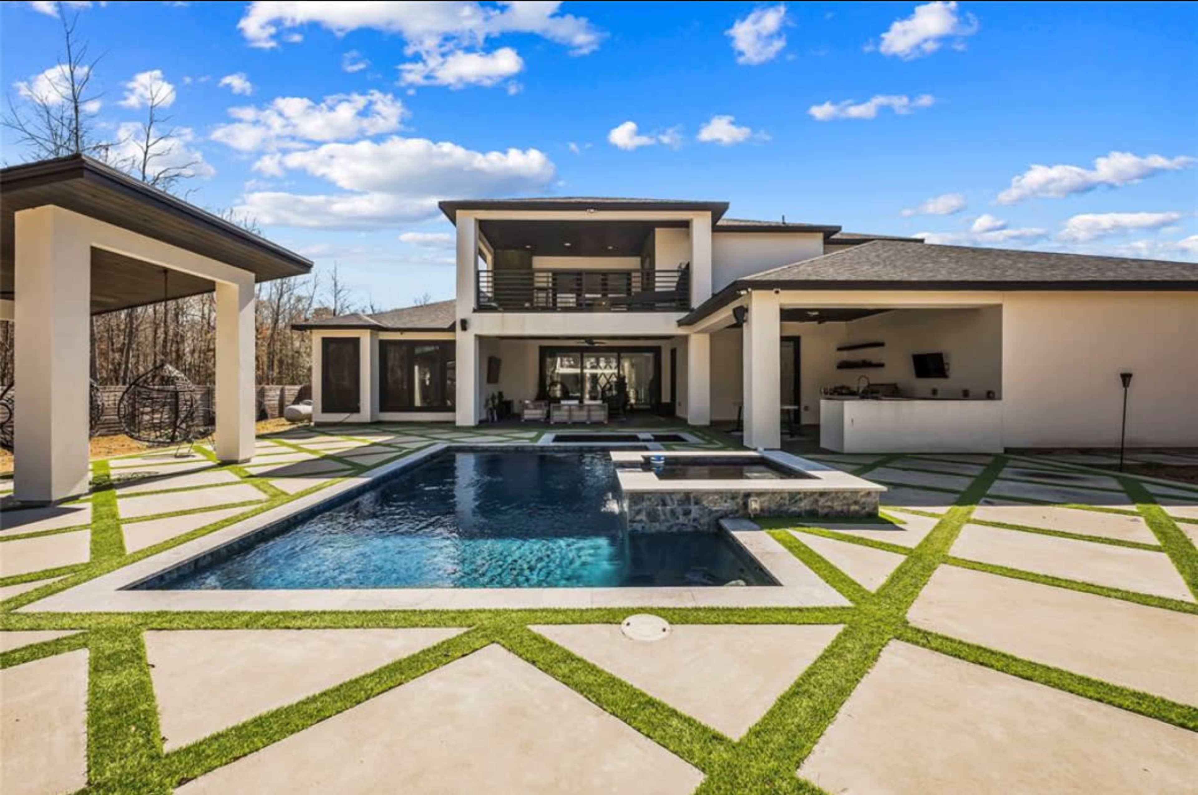 The image shows a modern house with a swimming pool surrounded by geometric patterns of grass and concrete, set against a backdrop of a clear blue sky with scattered clouds.