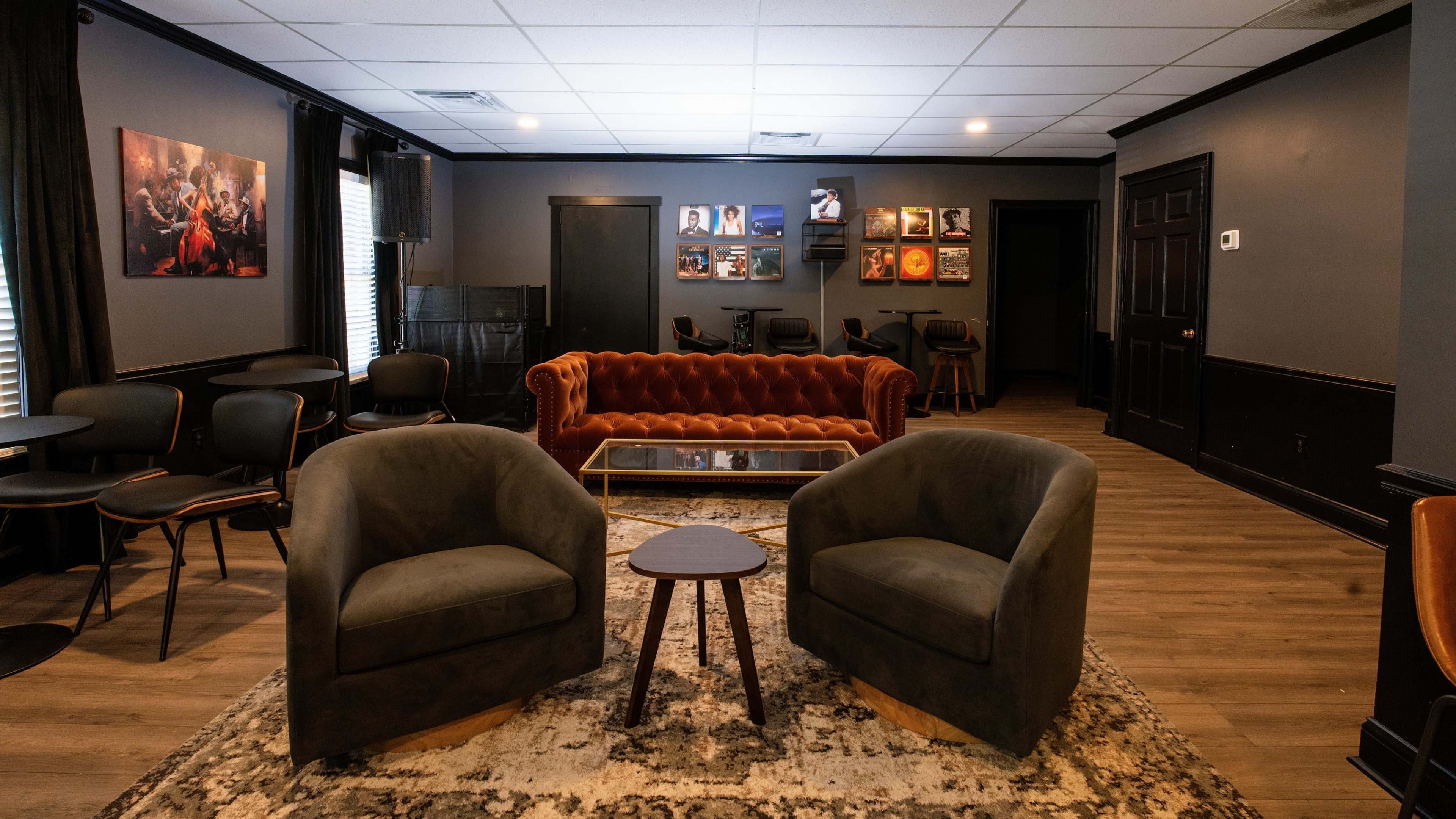 The image shows a cozy lounge area featuring a reddish-orange sofa, two gray armchairs, a small round table, and various framed artworks on the walls.