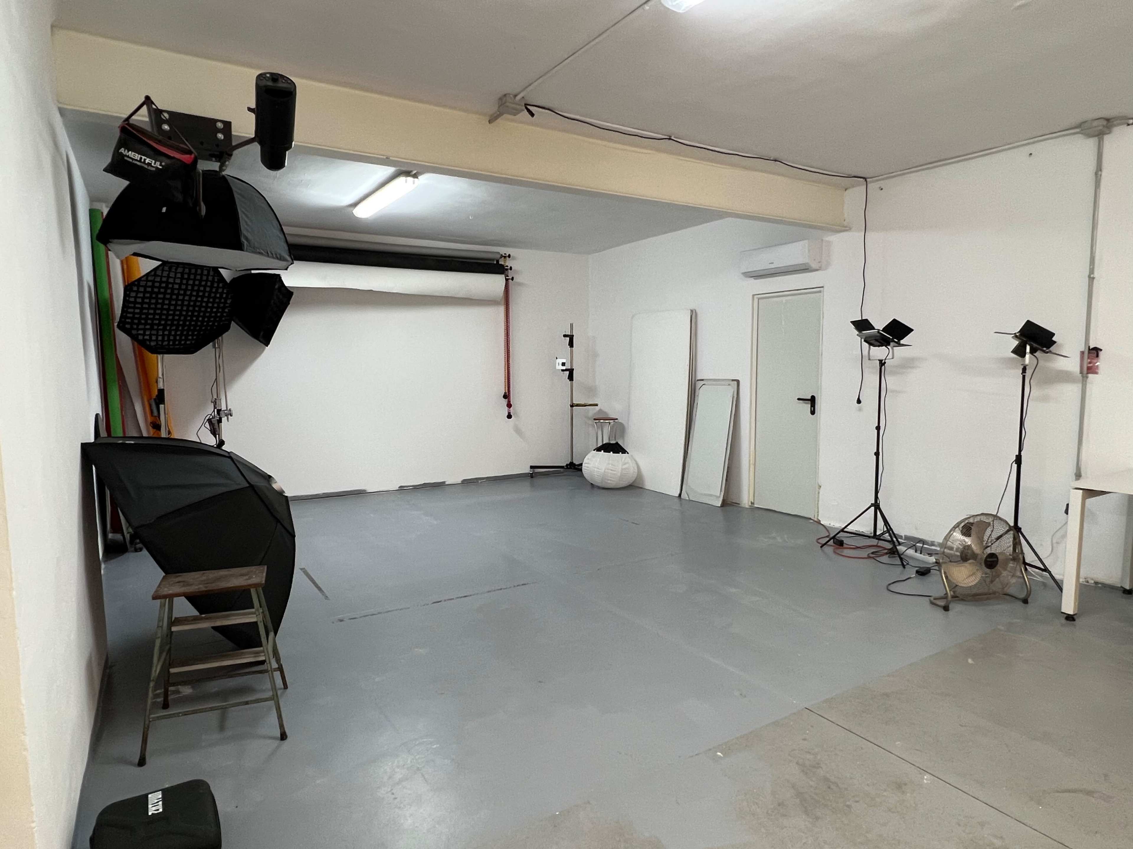 The image shows a photography studio with various lighting equipment, a fan, and a blank wall in the background.