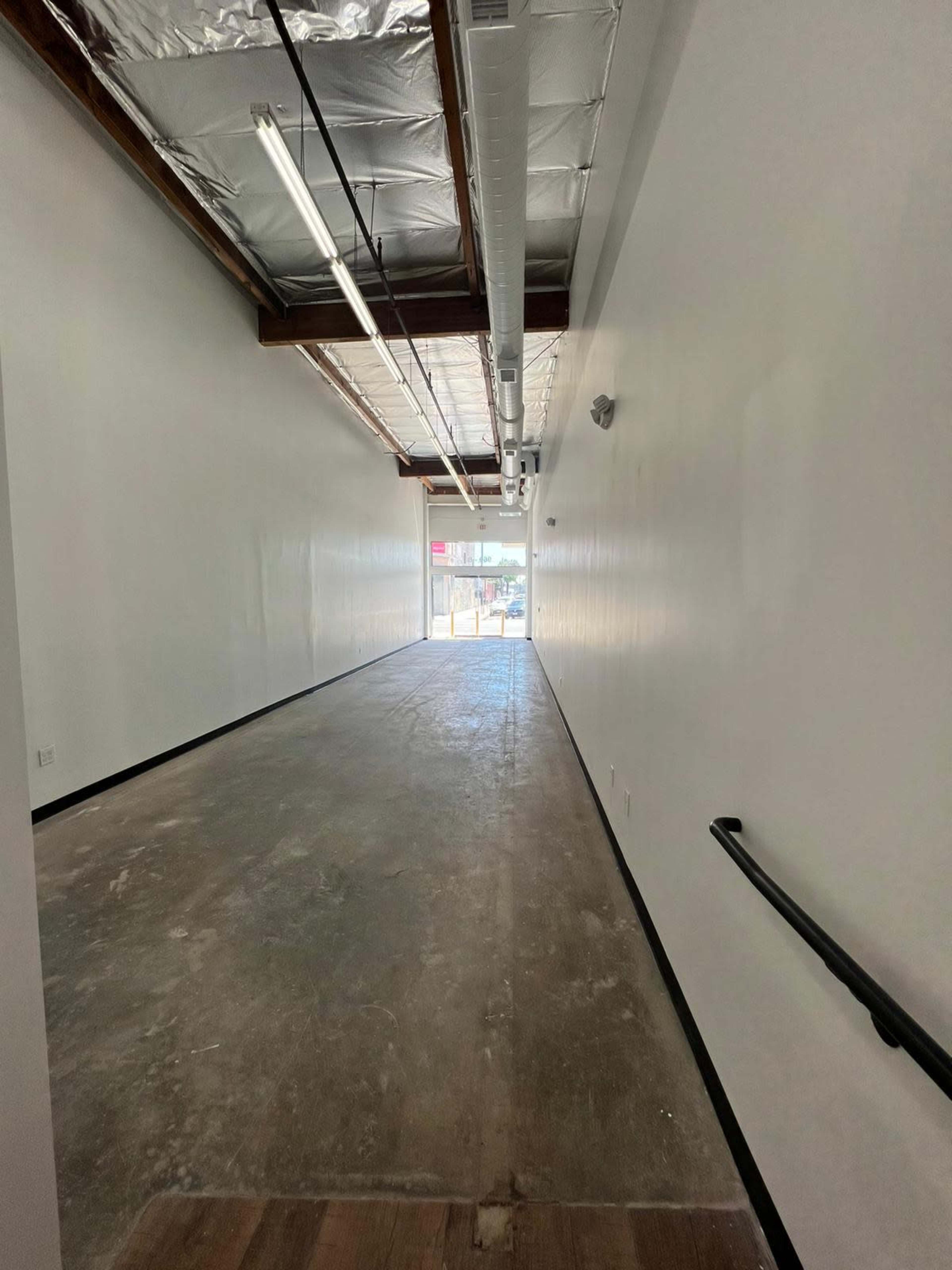 The image shows a long, empty corridor with concrete flooring and white walls, leading to a doorway with natural light at the end.