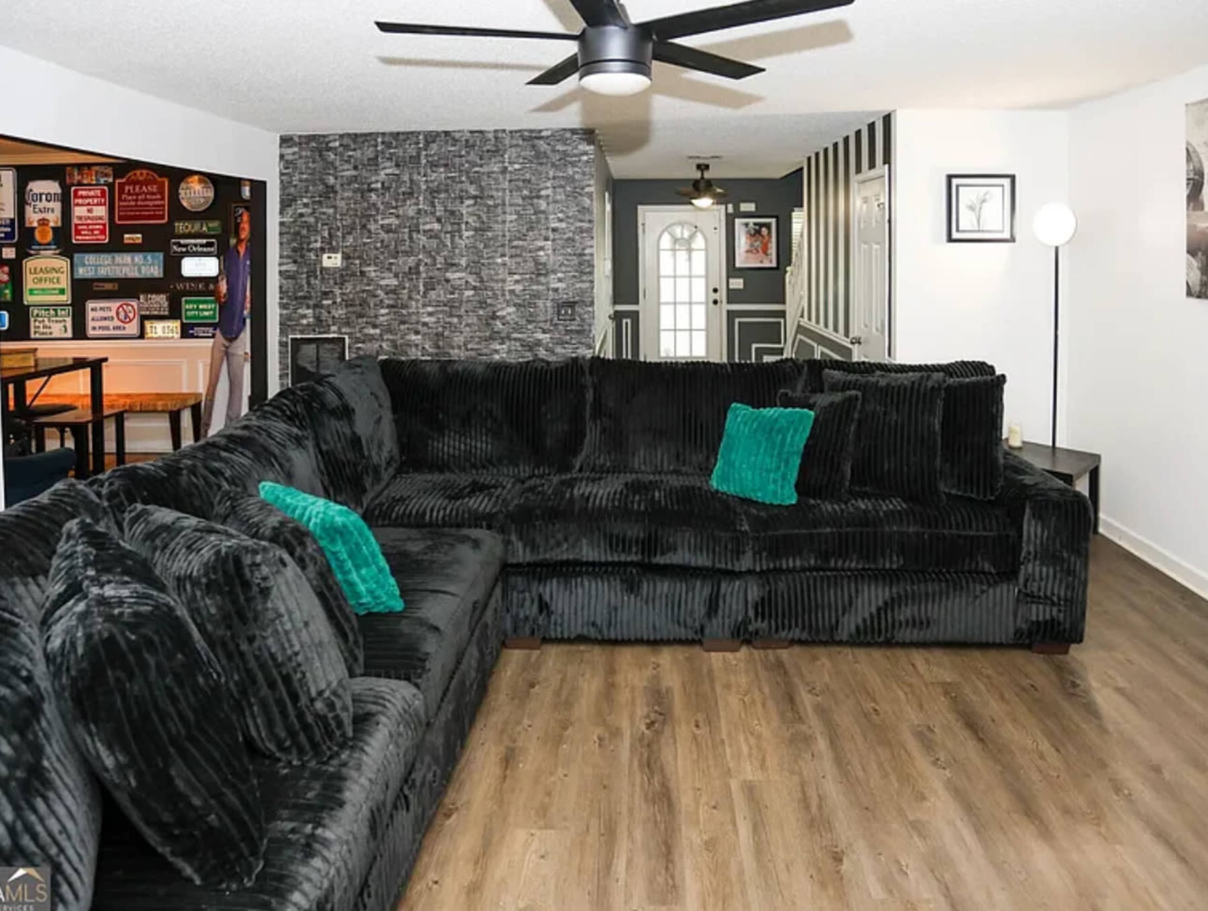 The living room features a large black sectional couch with teal pillows, hardwood flooring, and walls adorned with various signs and artwork.