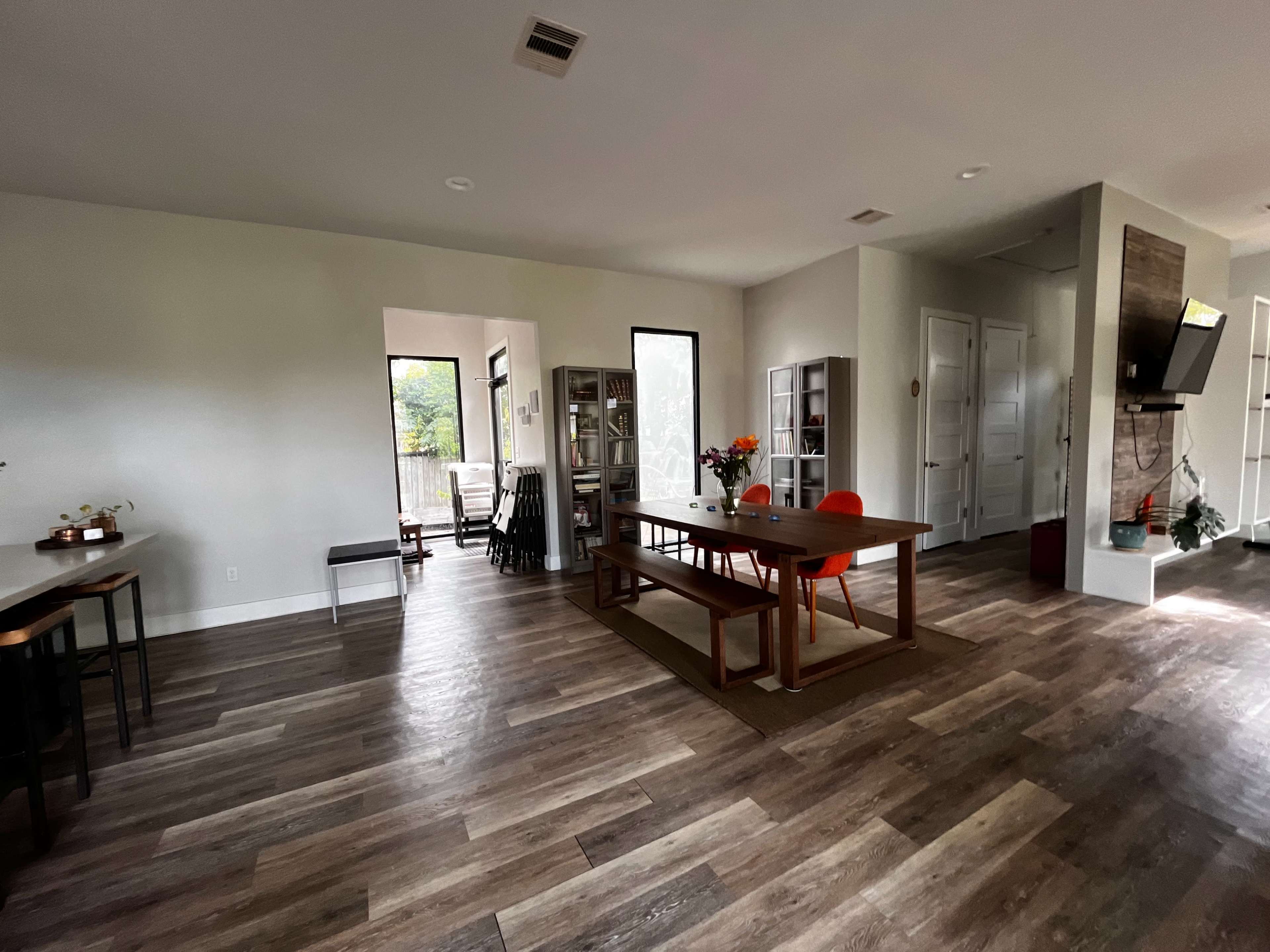 The image shows a spacious, lightly furnished interior with a dining table at the center, surrounded by wooden flooring and large windows allowing natural light.