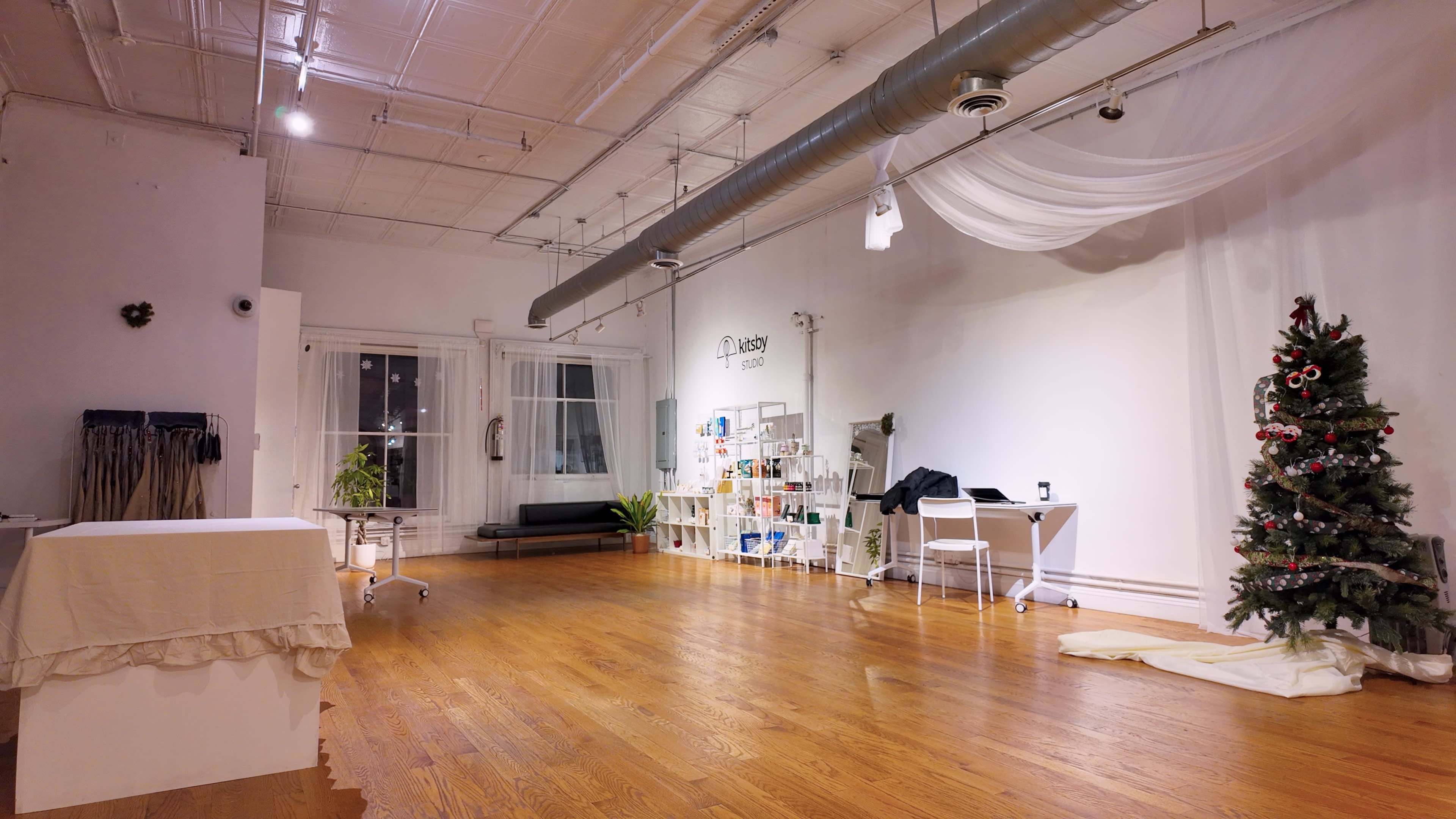 The spacious room features hardwood floors, a decorated Christmas tree, a white wall with a shelving unit, and minimal furniture, creating a bright and open atmosphere.