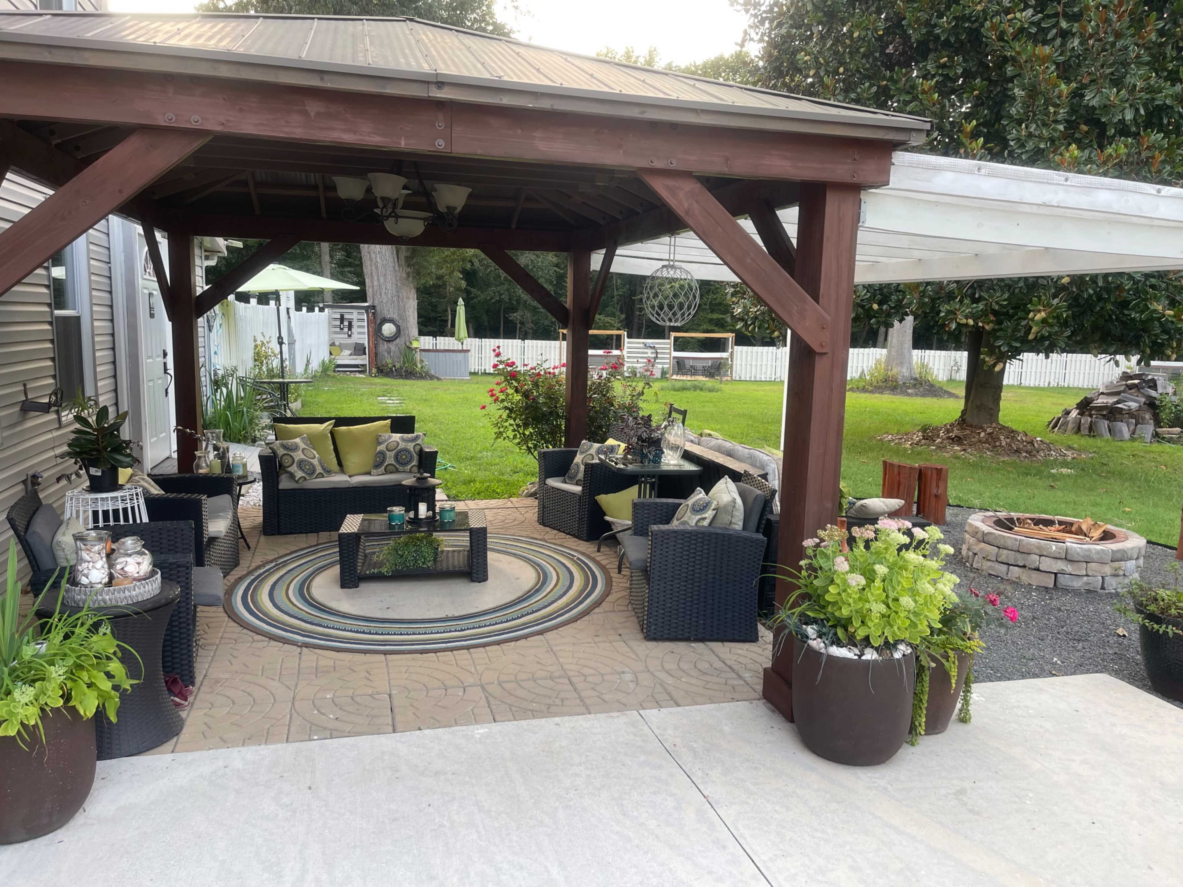 A covered patio features a seating area with dark wicker furniture and a circular rug, surrounded by potted plants and a fire pit on a stone patio.