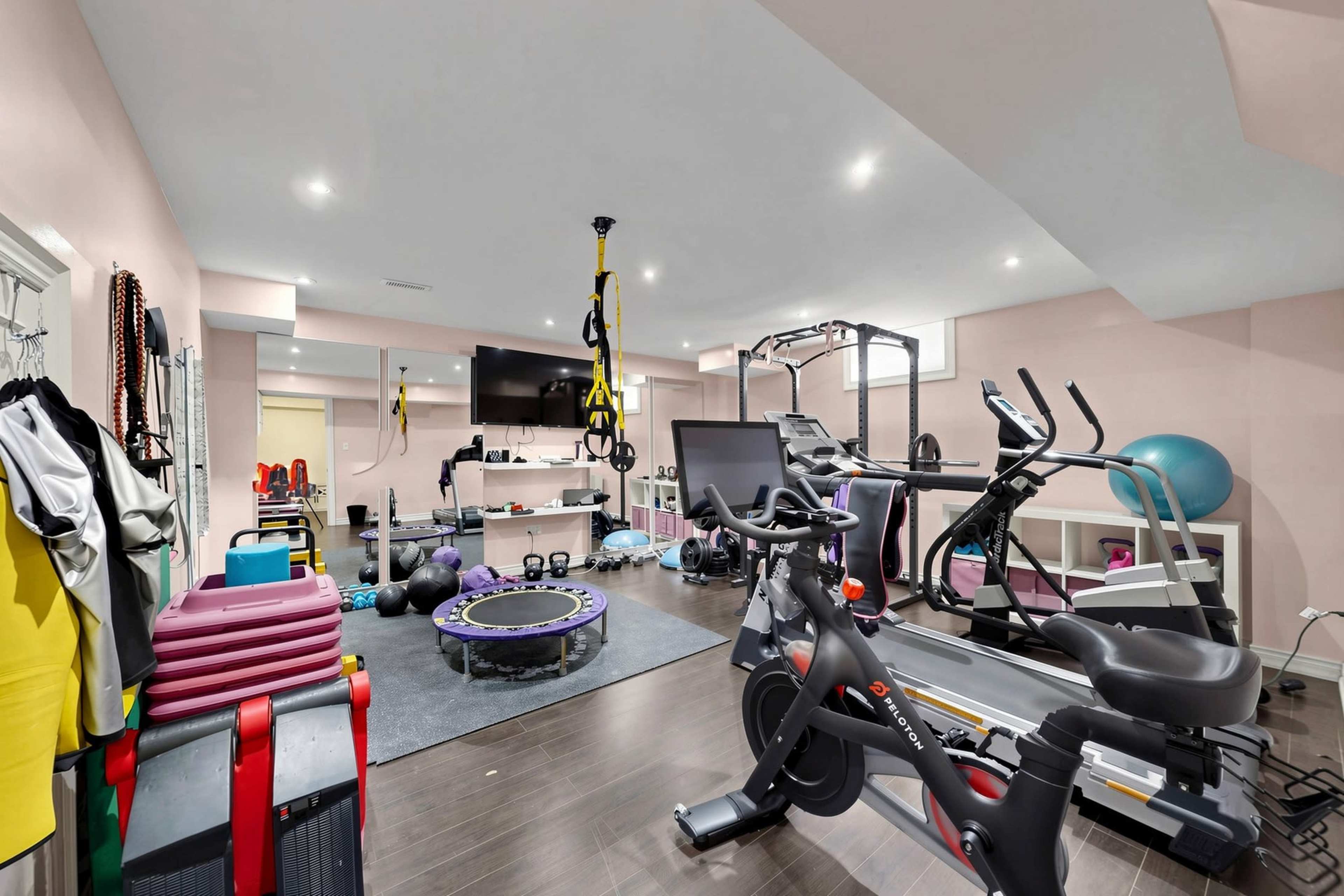 The image shows a home gym furnished with various exercise equipment, including cardio machines, weights, and fitness accessories, in a well-lit room.