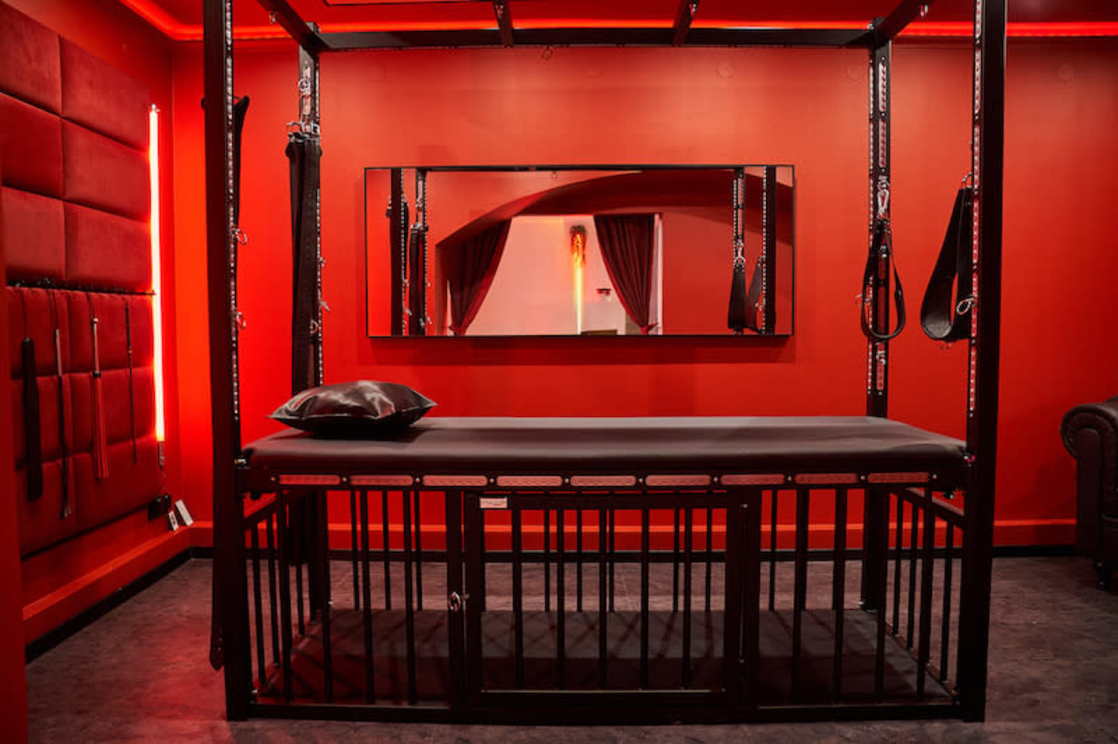 The image shows a red-themed room with a prominent metal frame structure, equipped with restraints and a black padded table in the center.