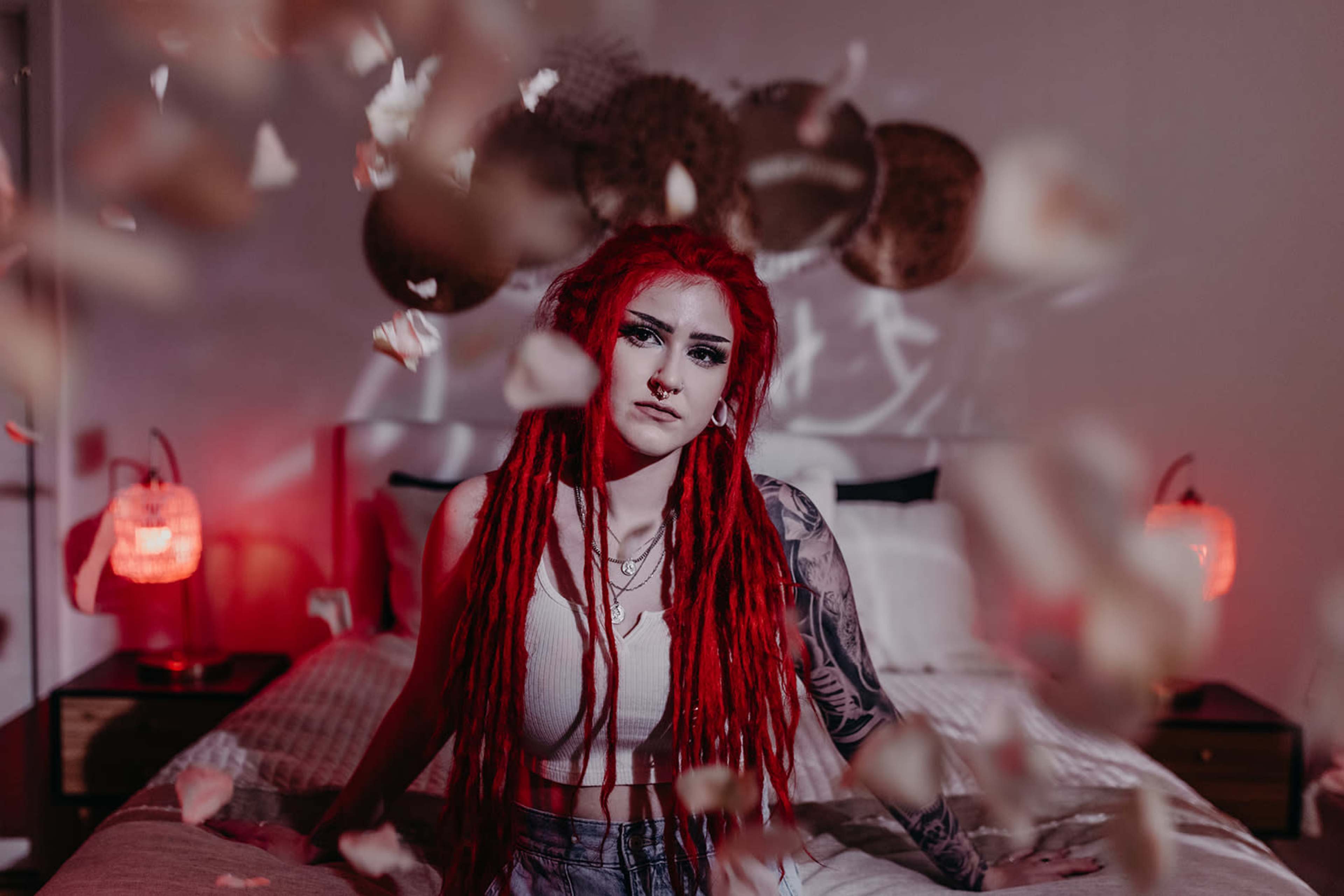 A person with red dreadlocks sits on a bed surrounded by floating rose petals in a softly lit room.