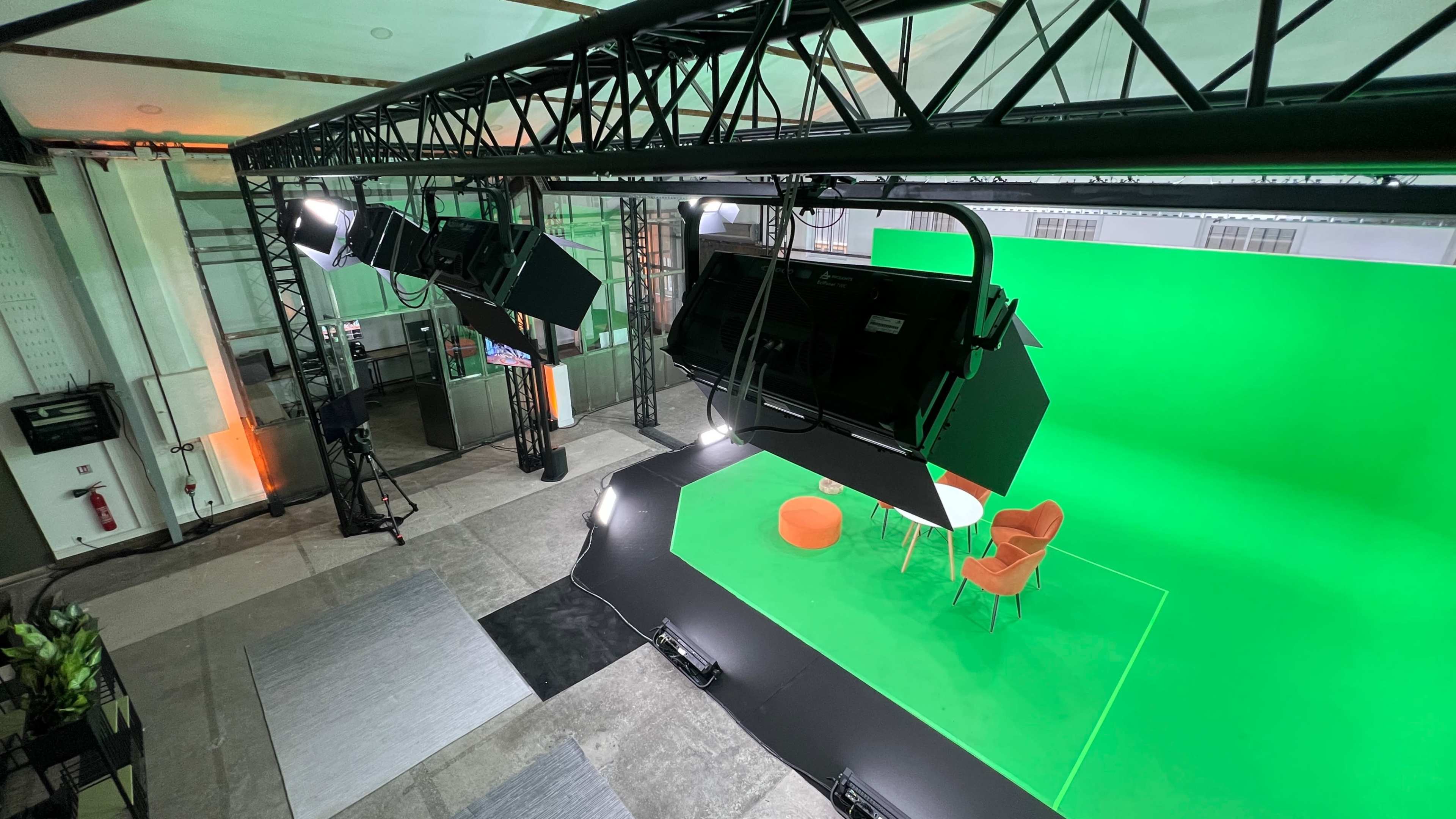 A studio setup with a green screen backdrop, two orange chairs, a table, and overhead lighting equipment.
