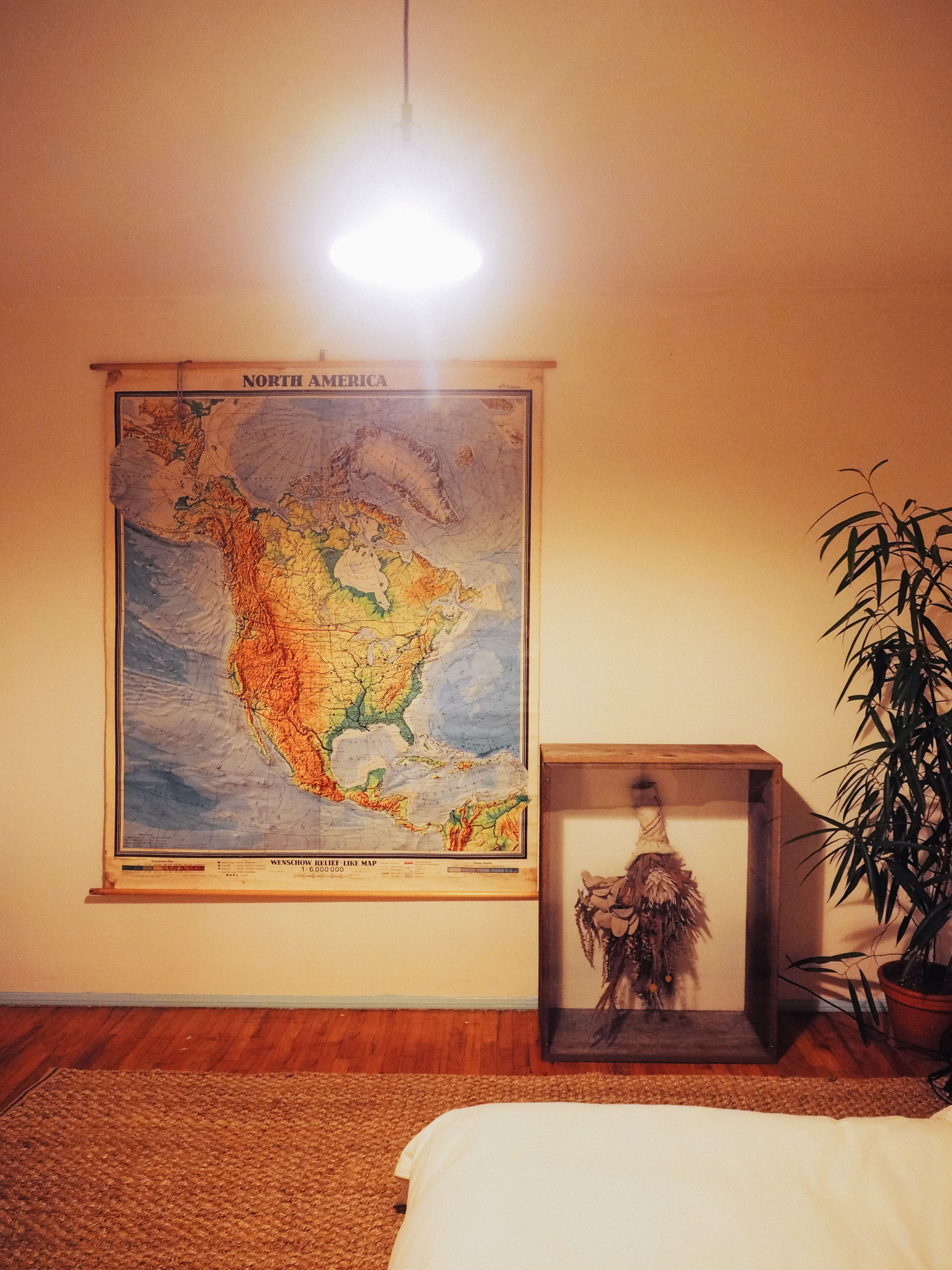A large map of North America hangs on the wall next to a glass display case containing a figure, with a plant in the corner and a bed covered by a white blanket.