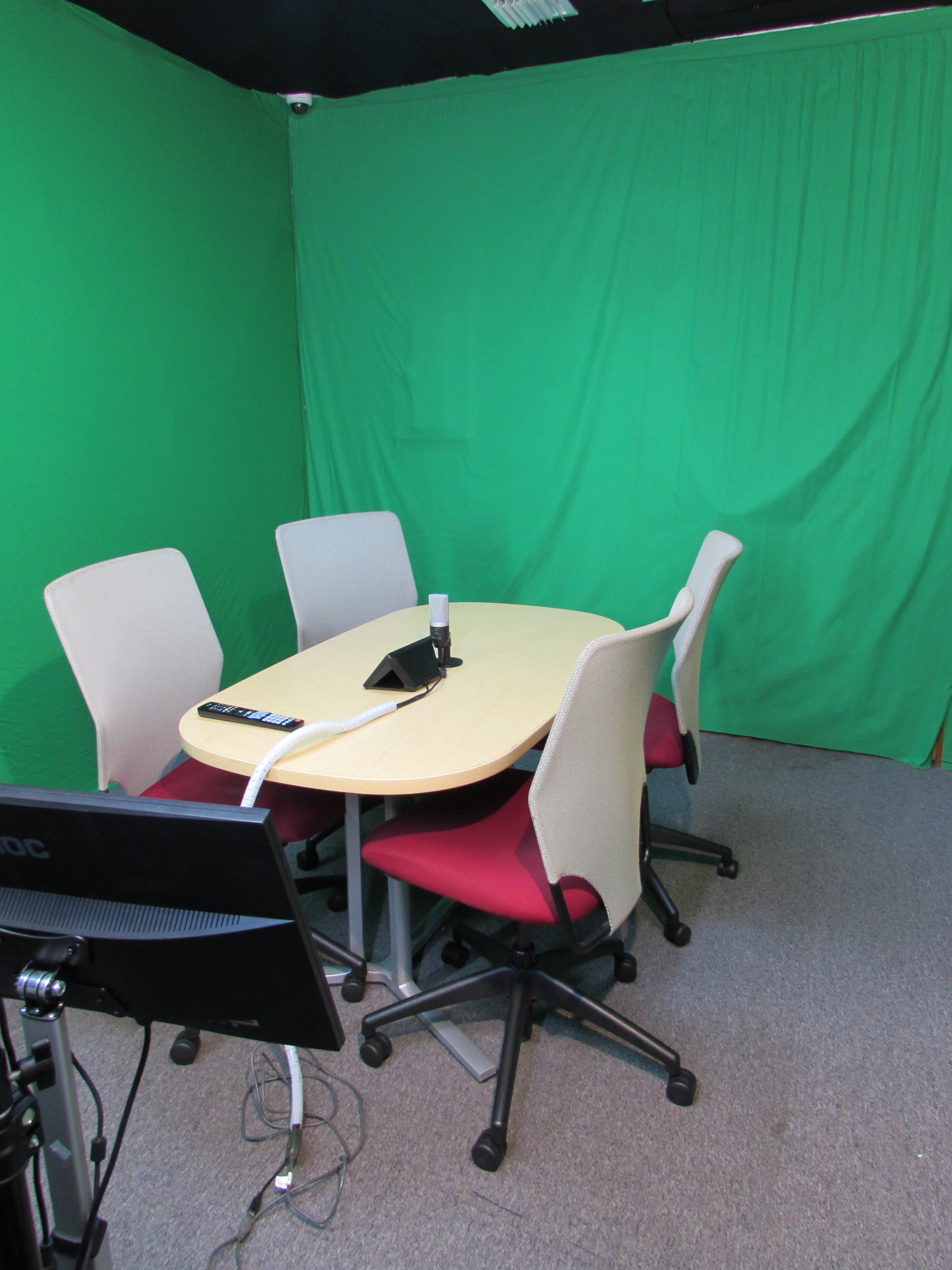 The image shows a small meeting room with a round table and four chairs in front of a green backdrop.
