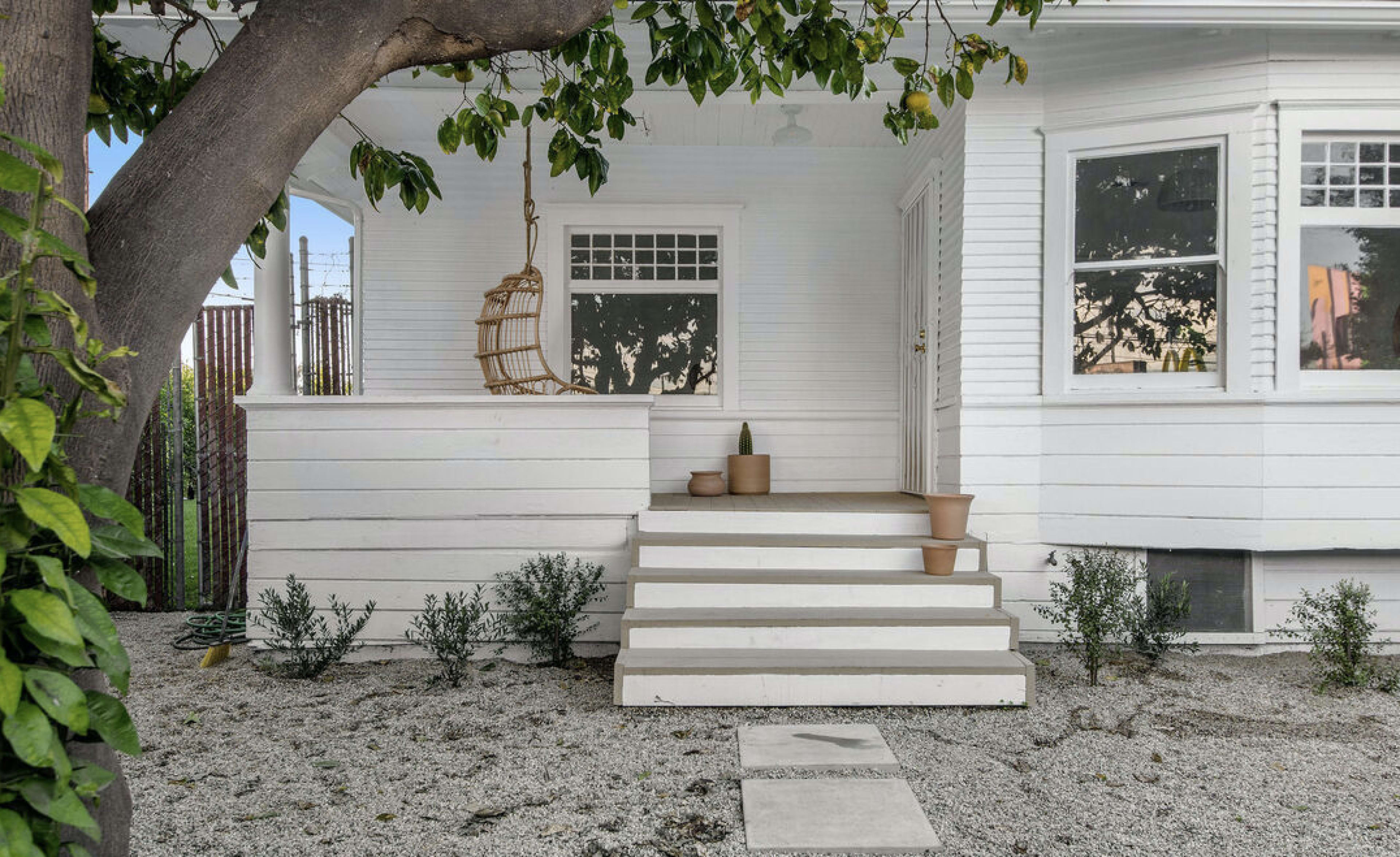 A white house with a front porch features a hanging chair and several potted plants on the steps.