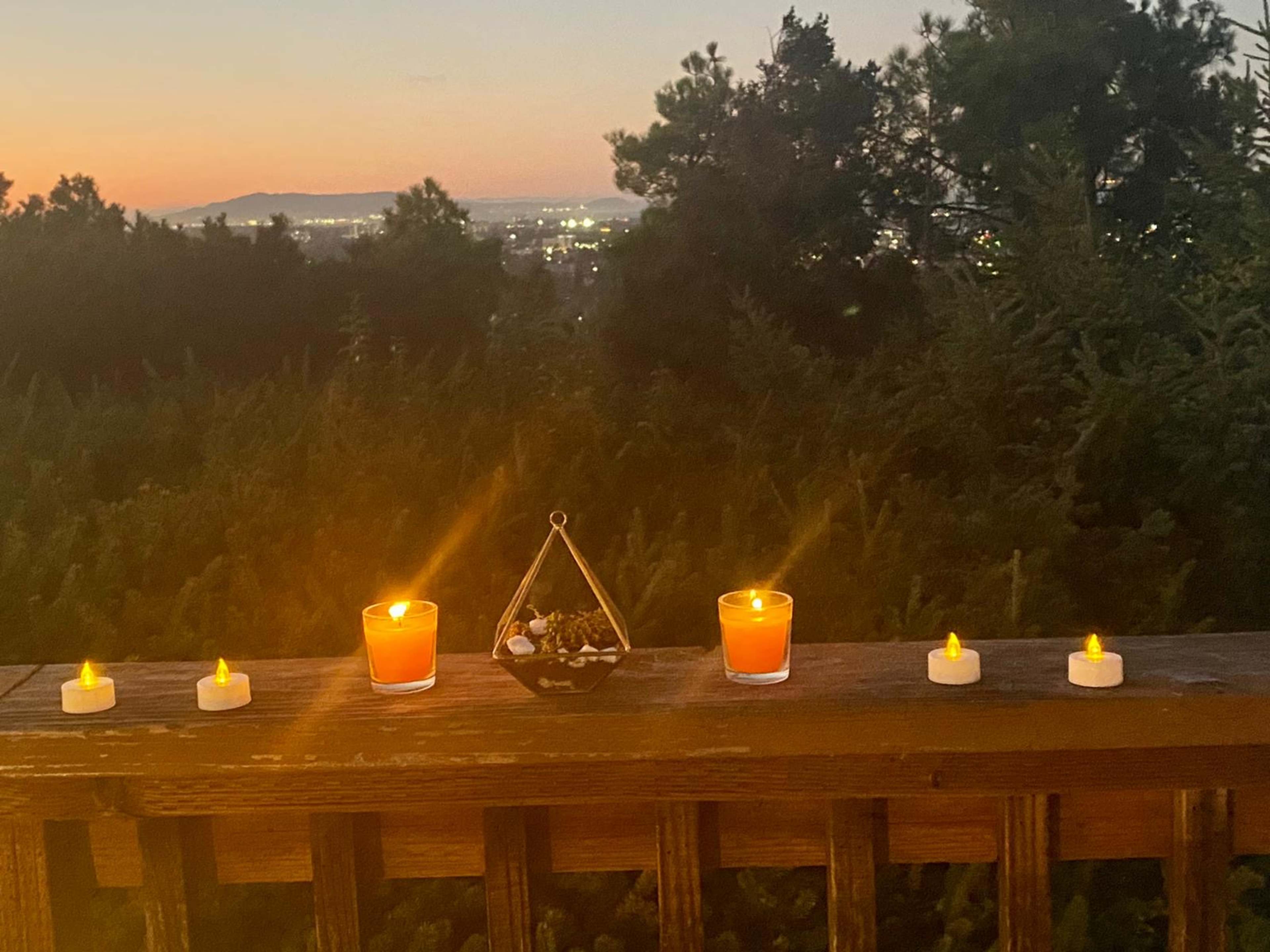 The scene shows a wooden railing adorned with candles and a decorative bowl, overlooking a landscape at twilight with mountains in the distance.