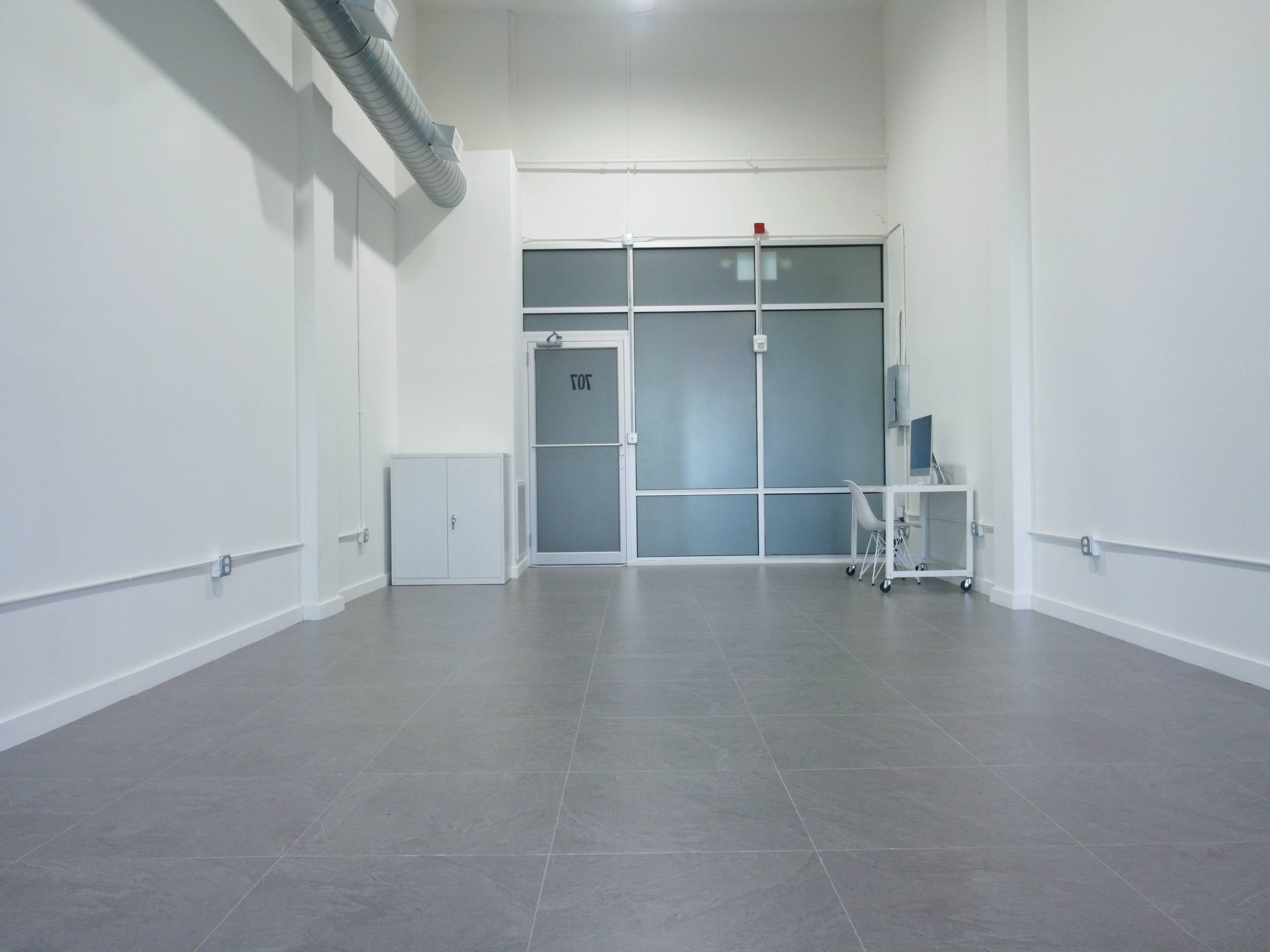 The image shows an empty room with gray tiled flooring, large front windows, and minimal furnishings, including a desk and chair near the entrance.