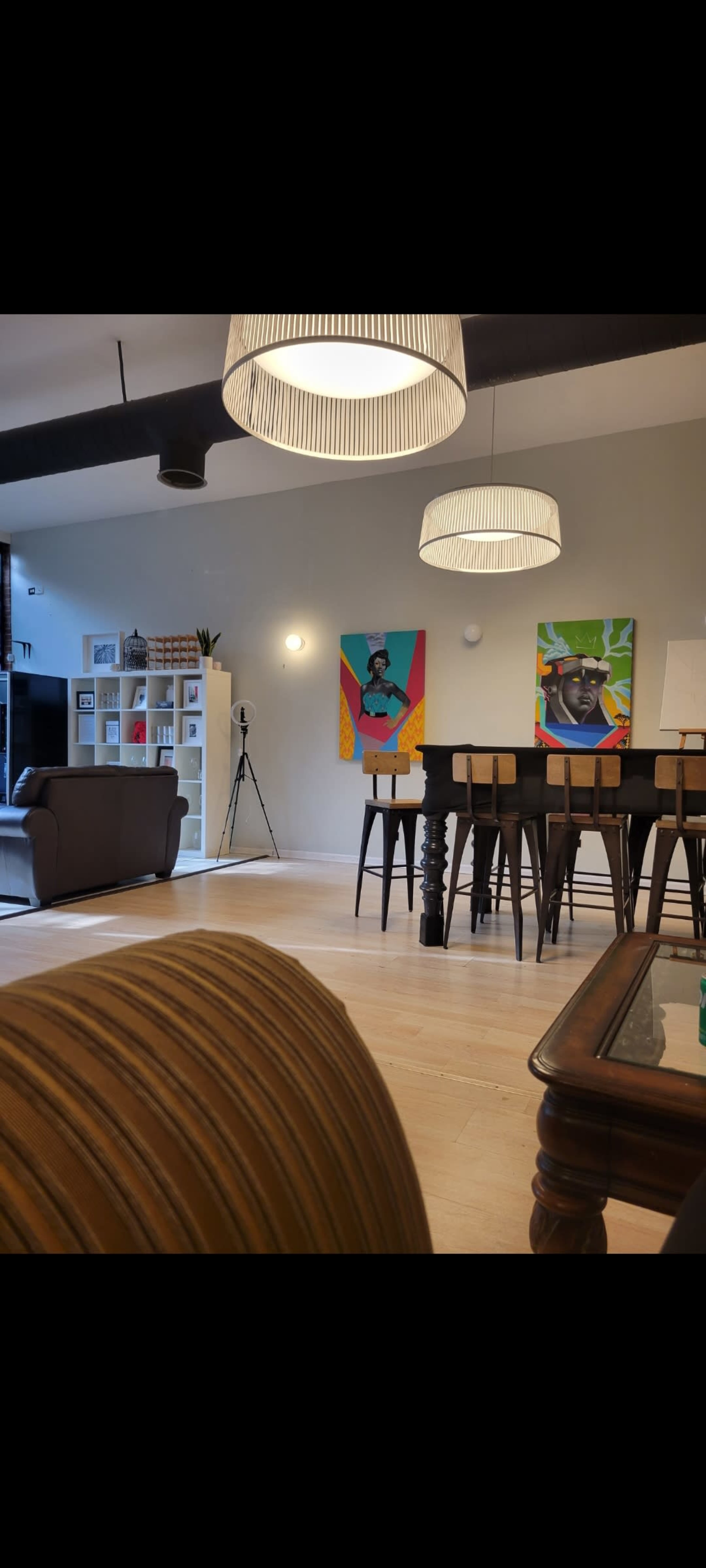 The image depicts a spacious room with two colorful wall paintings, a large table with stools, and a seating area featuring a couch.