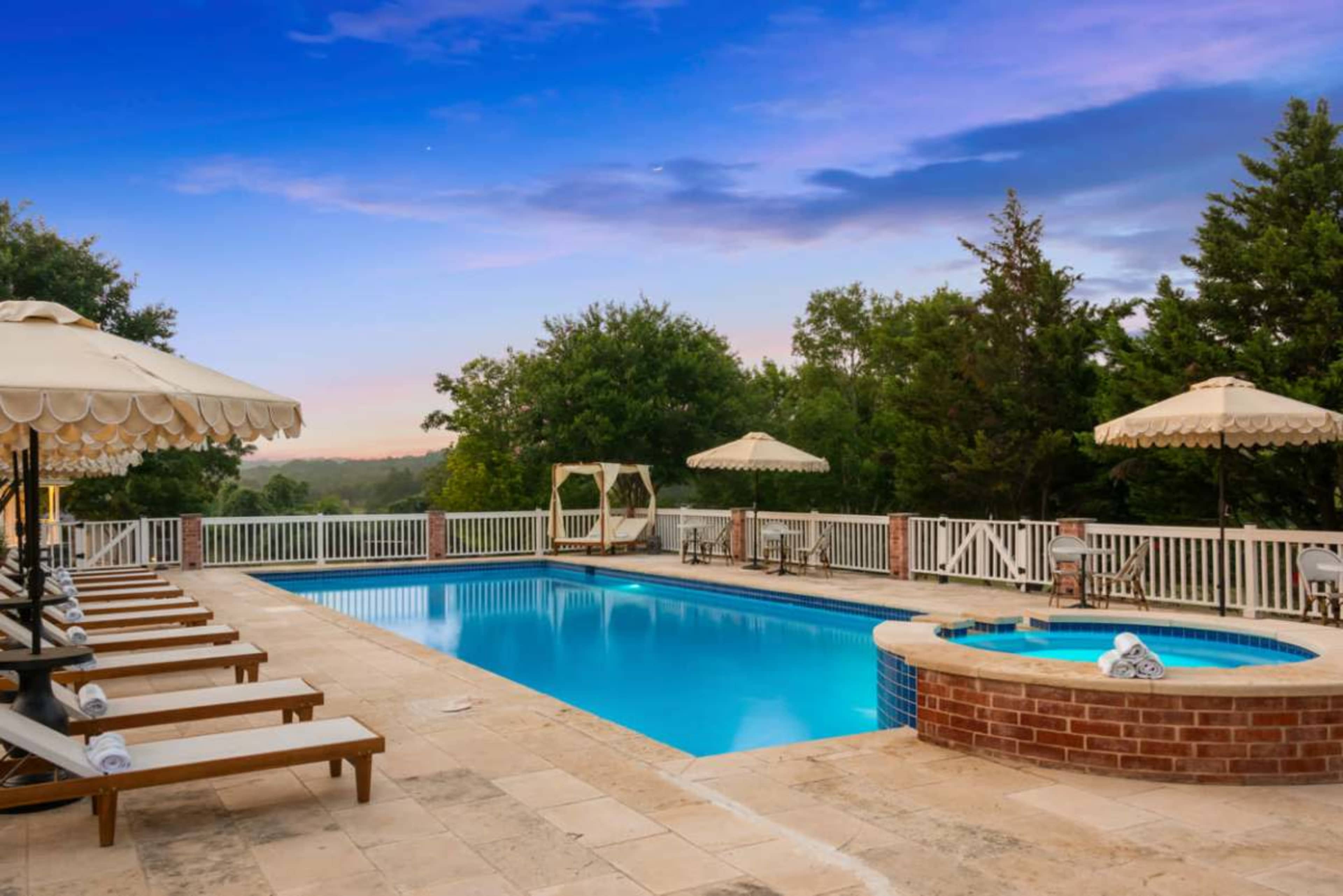 The image shows a well-kept outdoor pool area with sun loungers, umbrellas, and a hot tub surrounded by trees and a scenic sky.