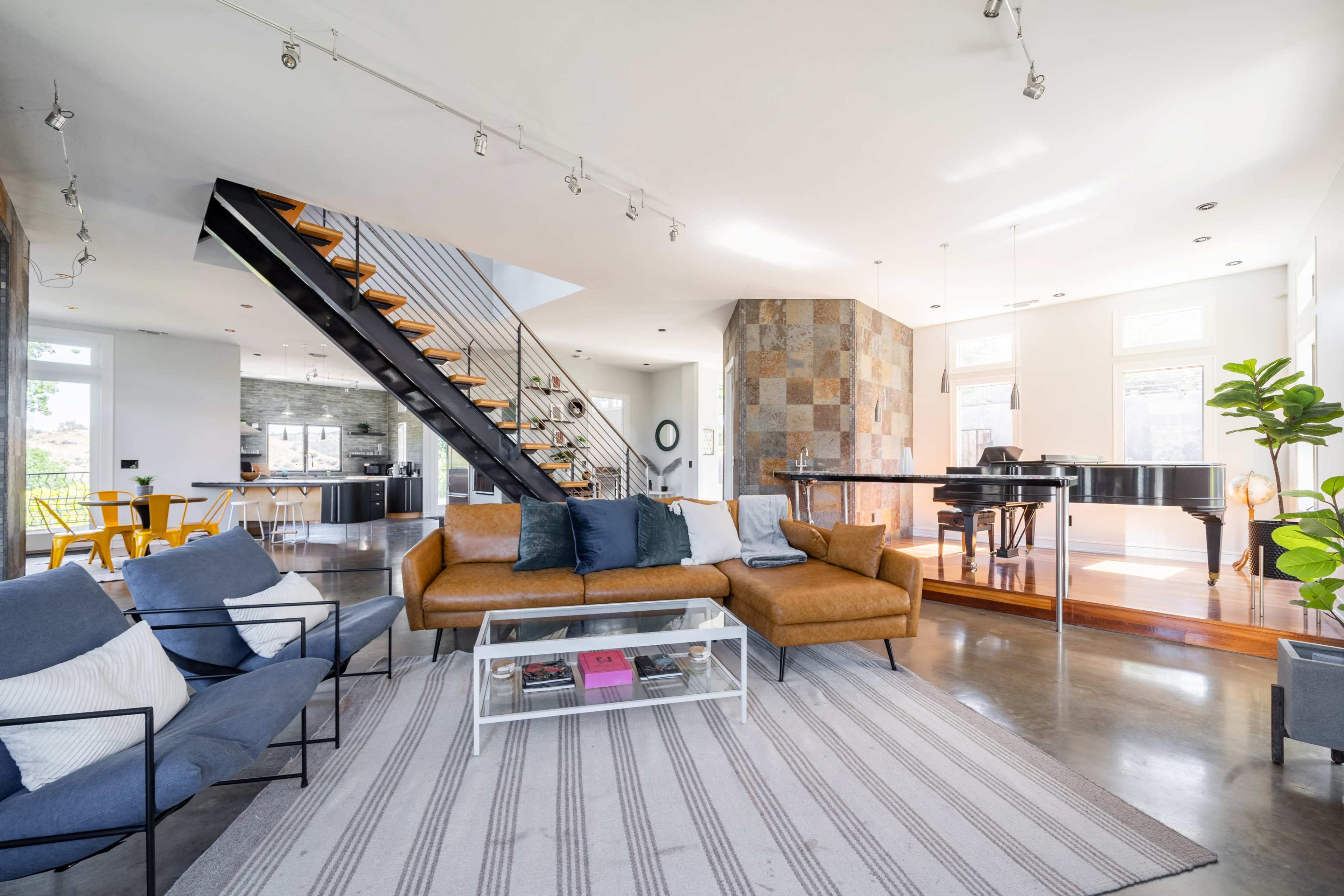 The image shows a modern, open-concept living space featuring a sectional sofa, coffee table, and a staircase leading to an upper level, with a grand piano positioned in a well-lit area.