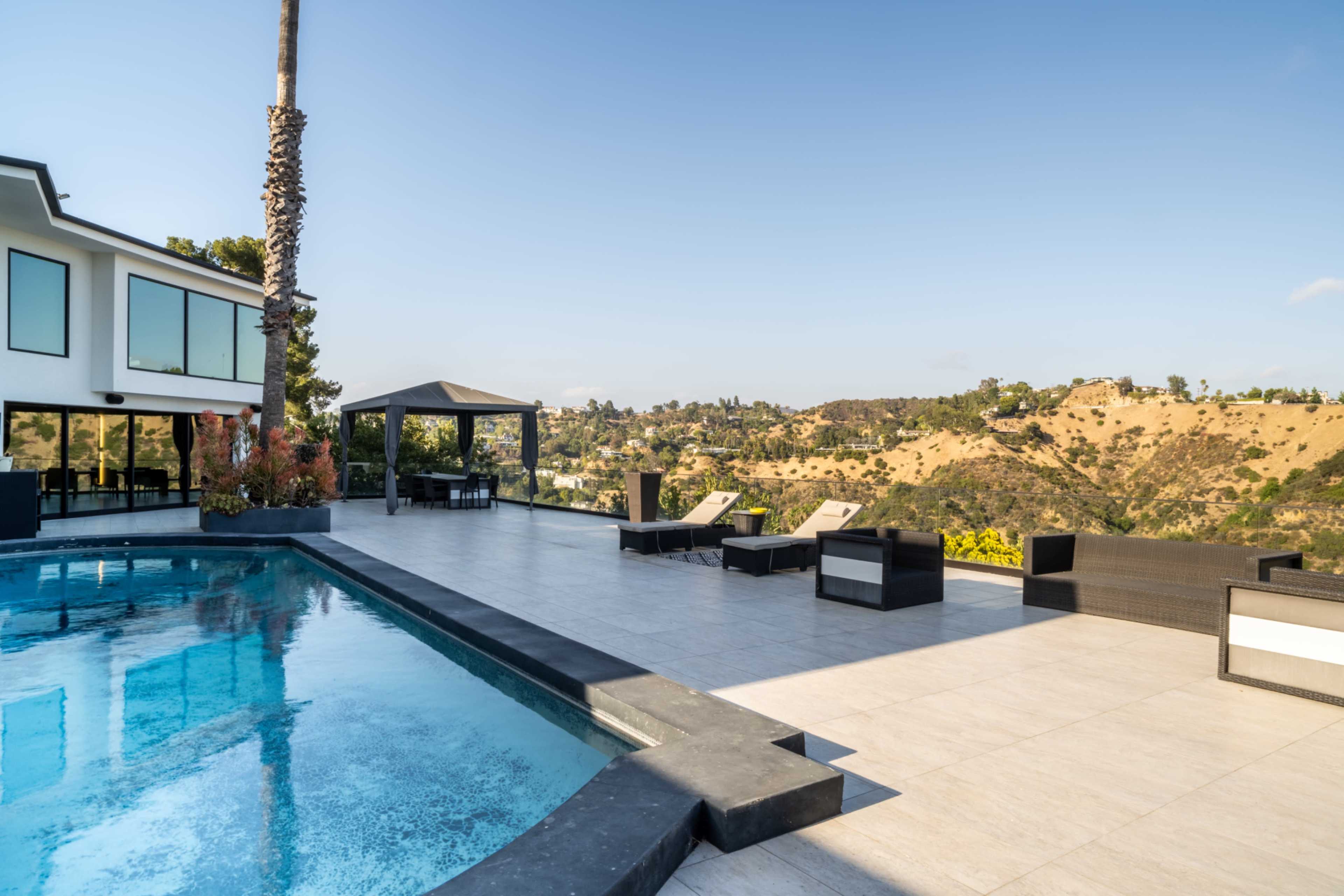 The image shows a modern outdoor patio with a swimming pool, lounge chairs, and a gazebo overlooking a hilly landscape.