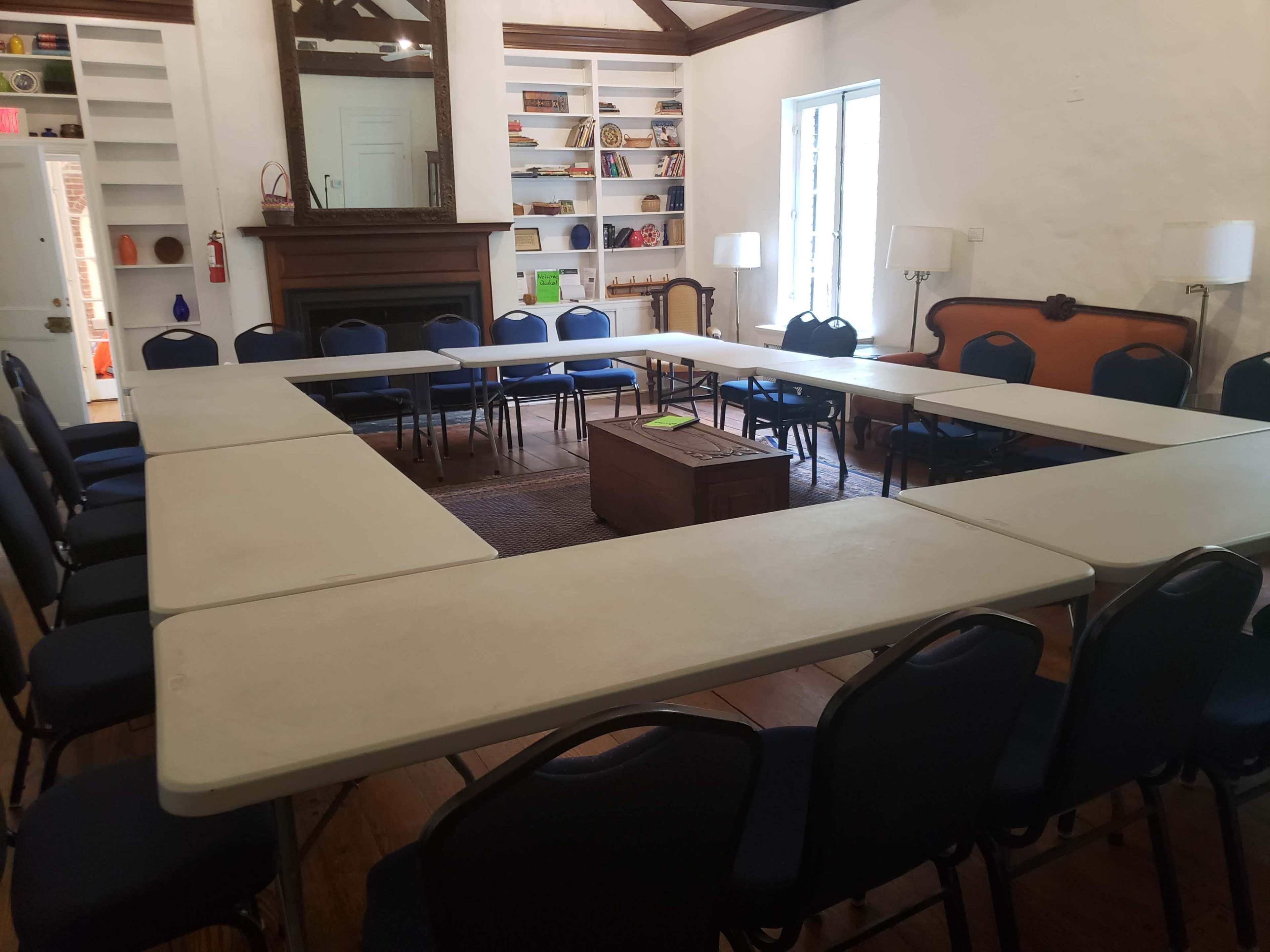 A meeting room features a rectangular arrangement of tables surrounded by chairs, with shelves and a fireplace in the background.