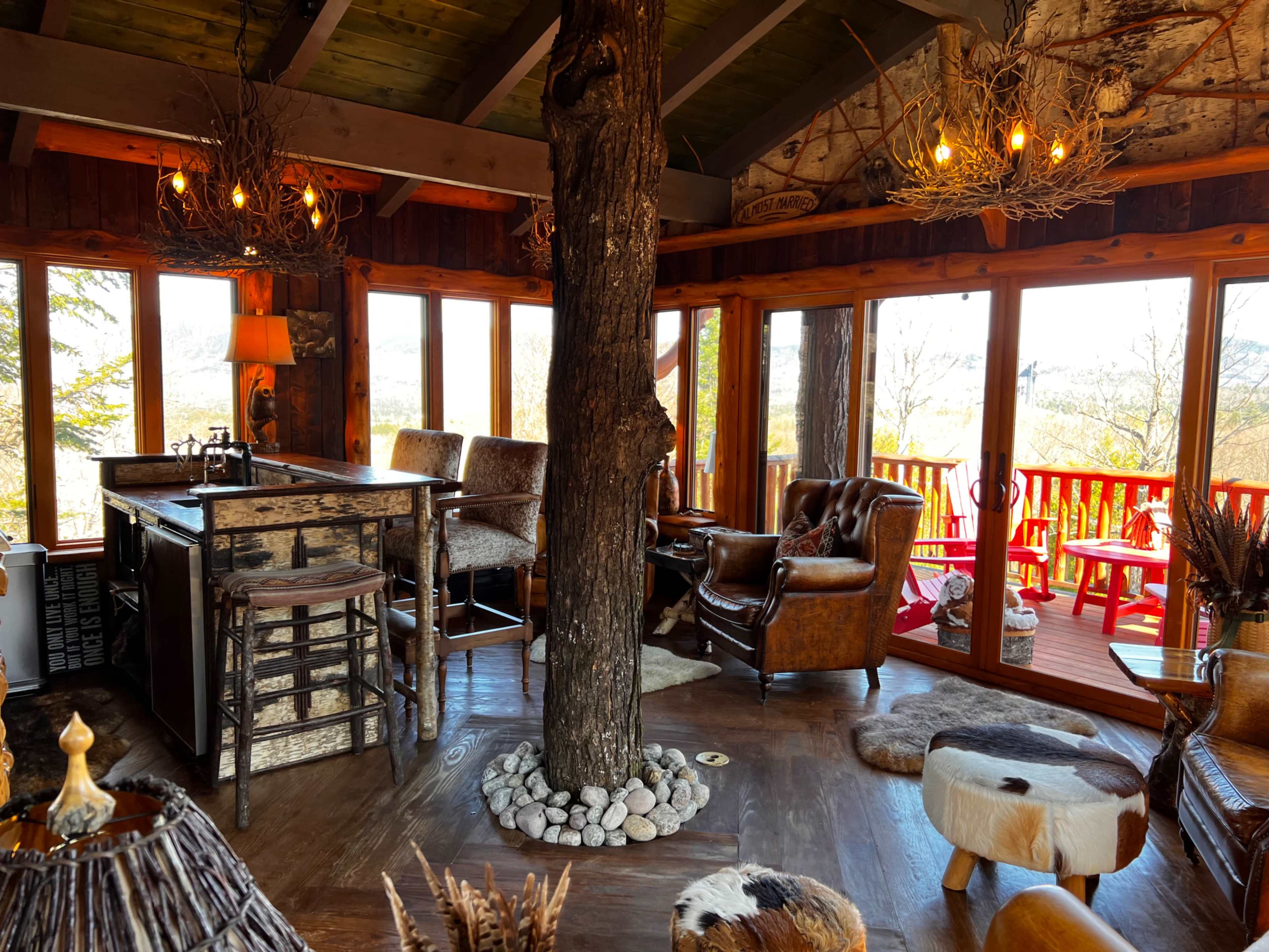 A rustic interior of a cabin with wooden walls and a bar area, featuring a large tree trunk as a central support, surrounded by comfortable seating and large windows that offer a view of the outdoors.