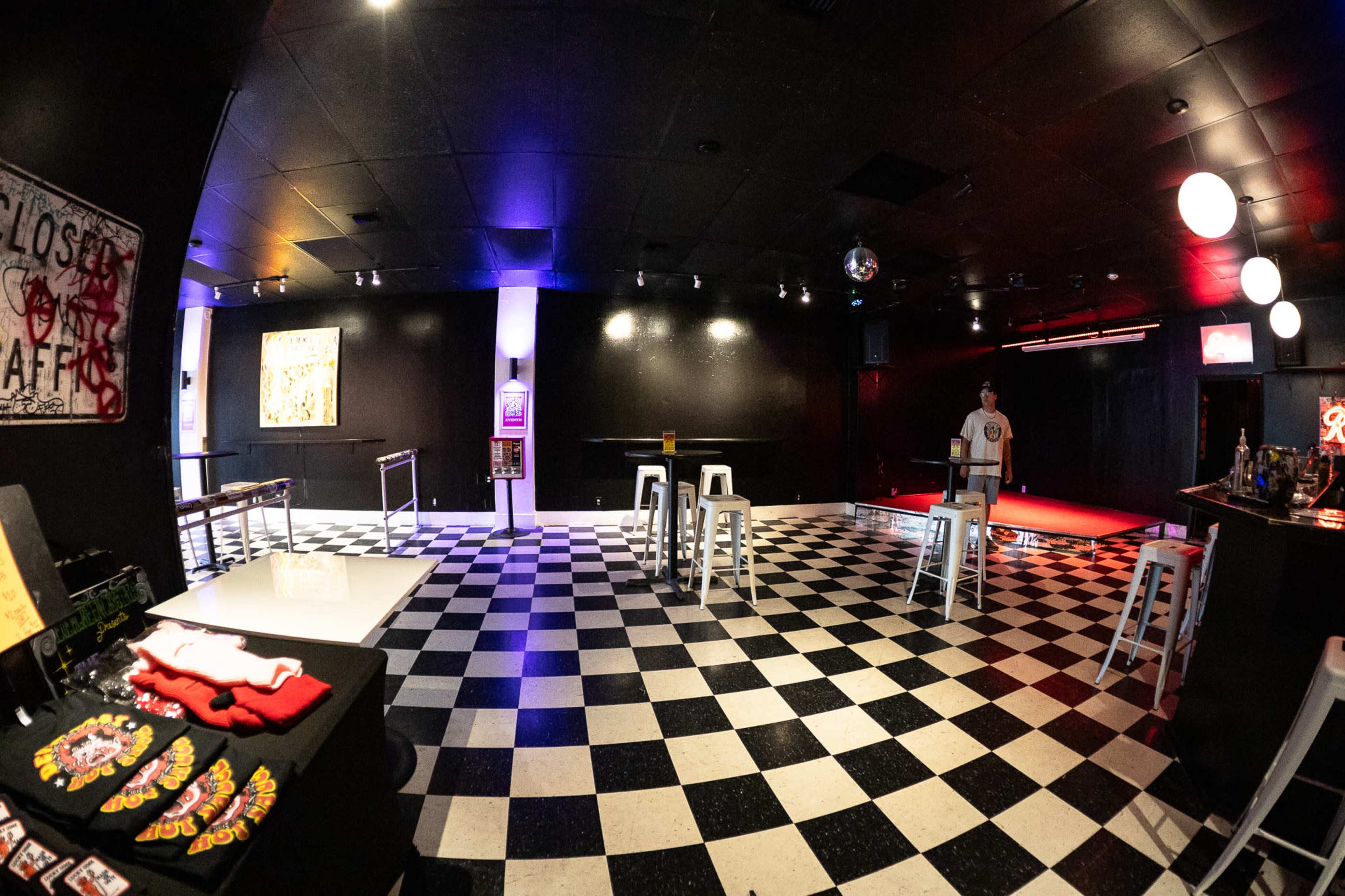 The image shows an empty black-and-white checkered room with bar stools and a small stage area, illuminated by colorful lights.