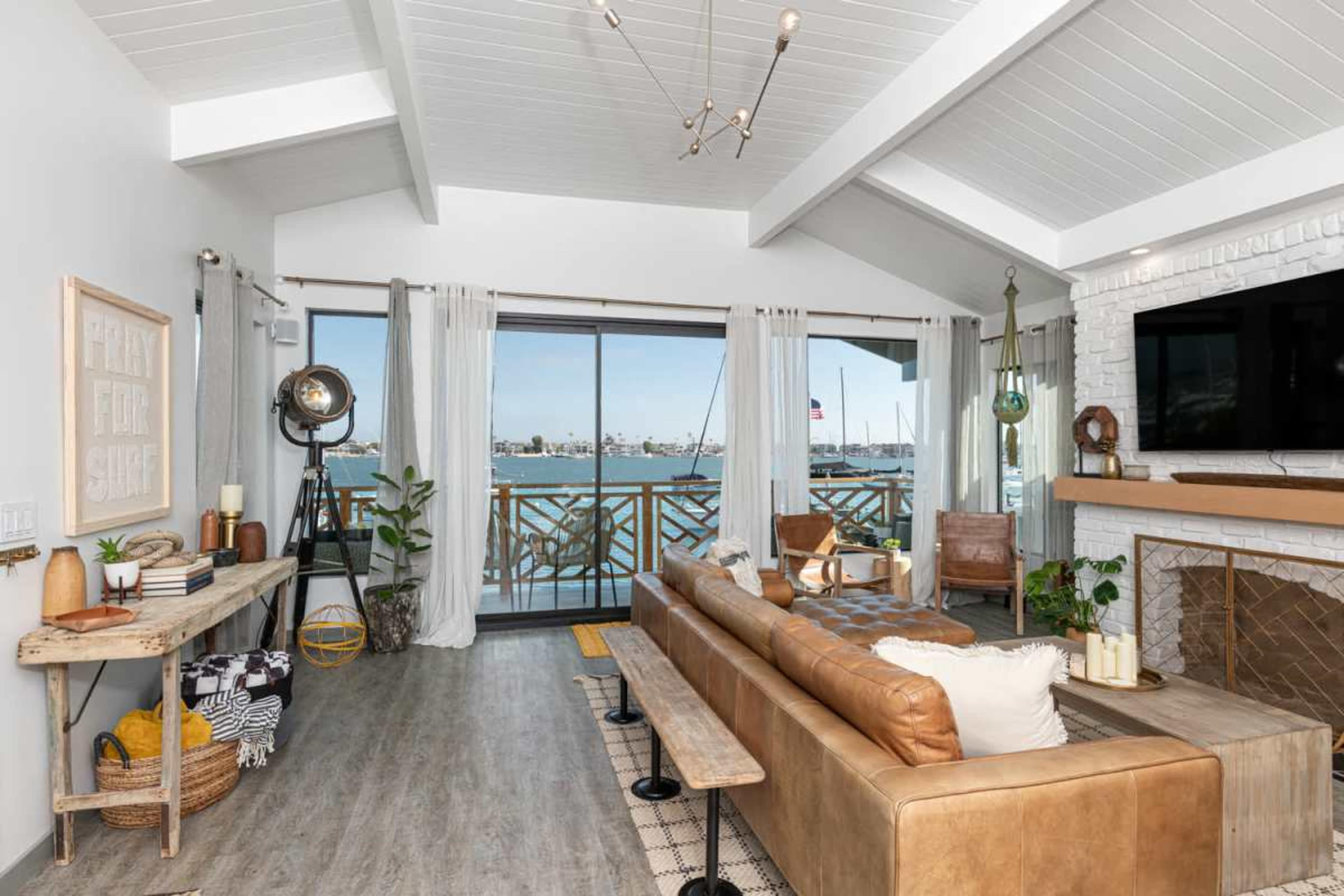 A spacious living room features a large window overlooking a harbor, with a brown leather sofa, wooden furniture, and coastal-inspired decor.