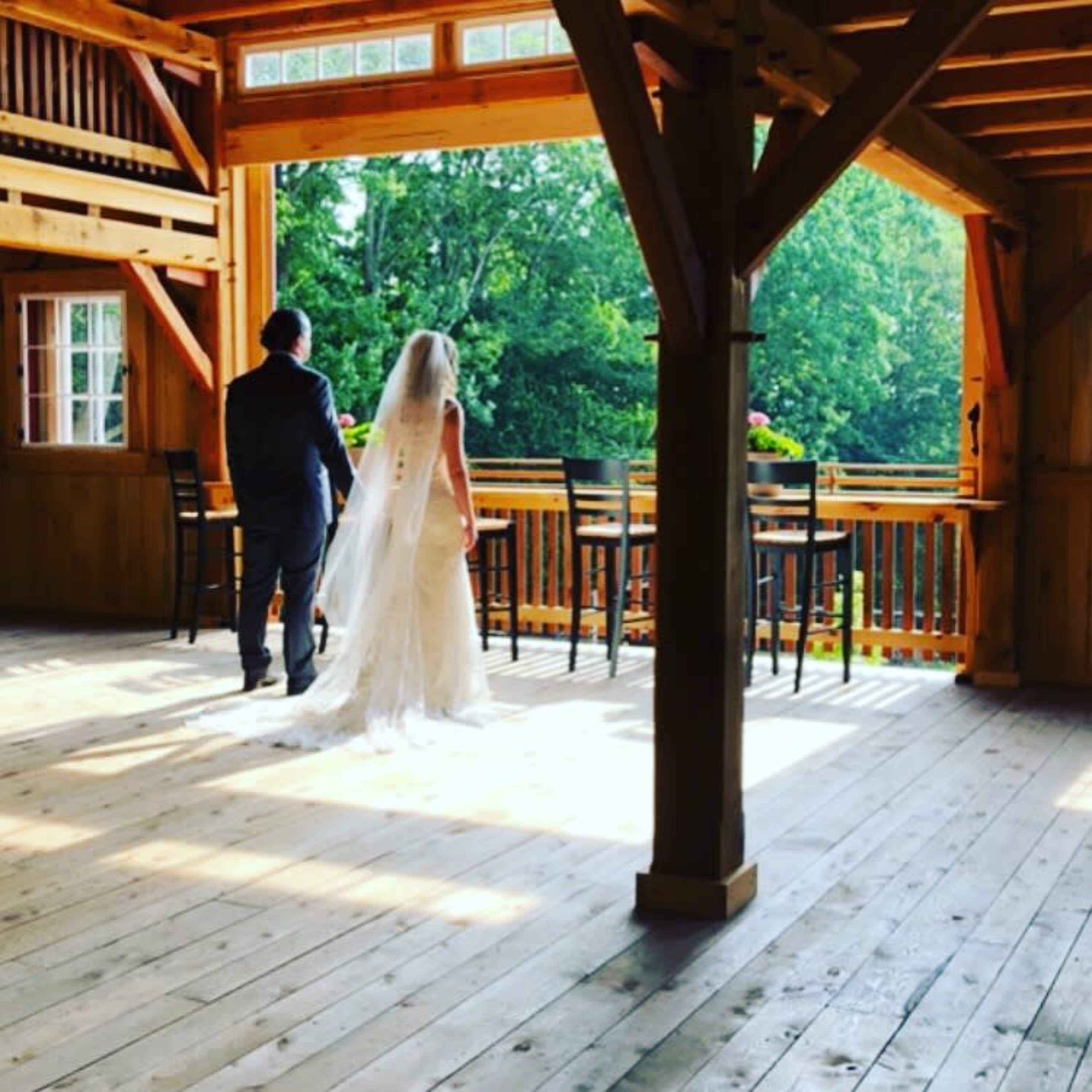A bride and groom walk together on a wooden floor inside a cabin with large windows overlooking a forested area.