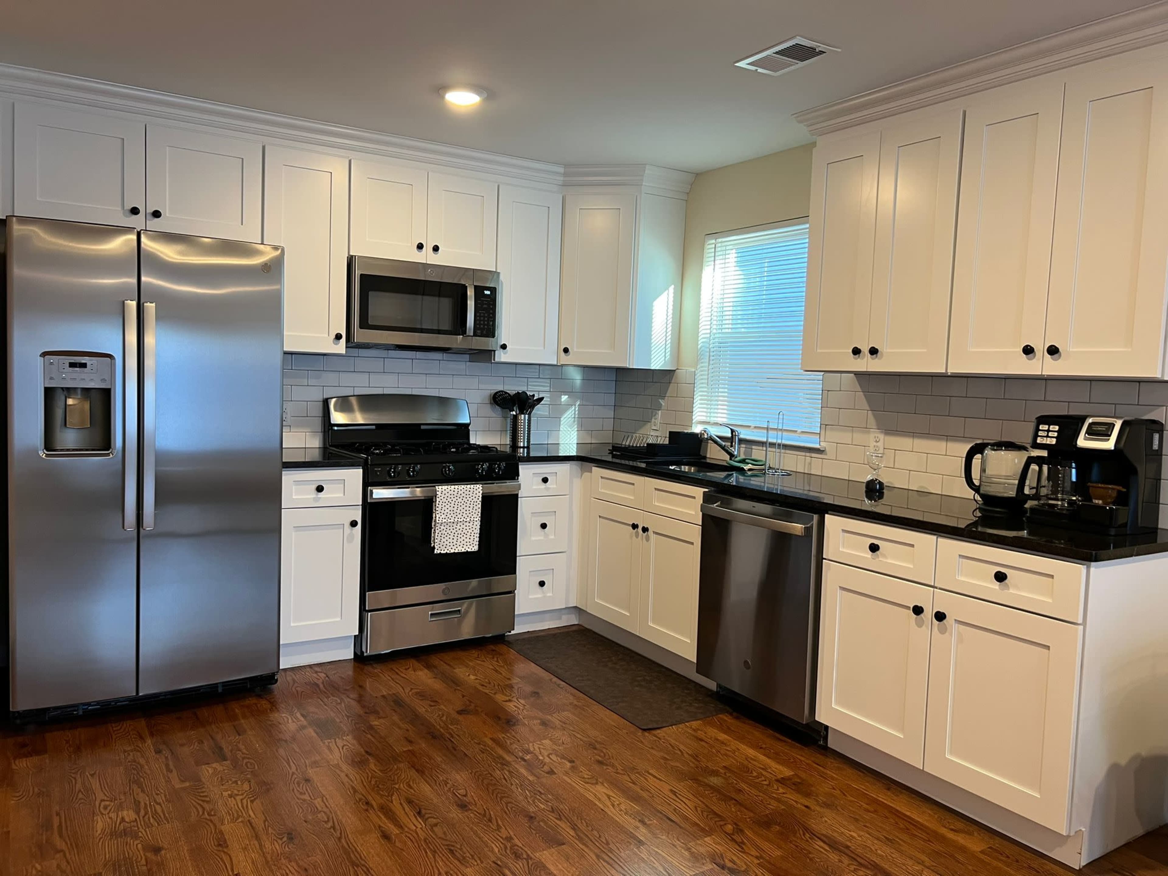A modern kitchen features stainless steel appliances, white cabinetry, and dark countertops, with a wooden floor and ample natural light.