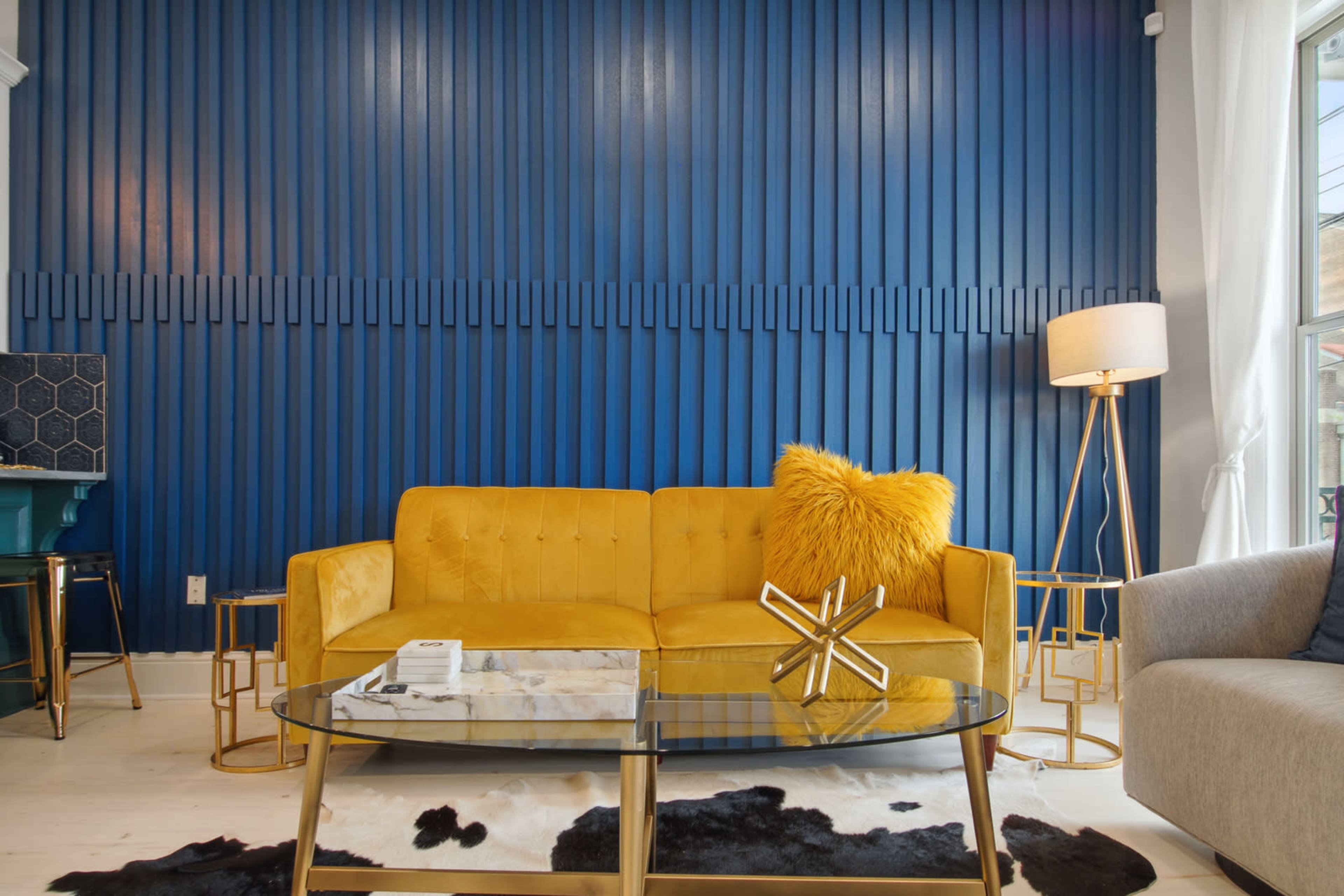 A yellow sofa with a fluffy pillow is positioned in front of a blue paneled wall, accompanied by a glass coffee table and a lamp, with a second lighter sofa nearby.