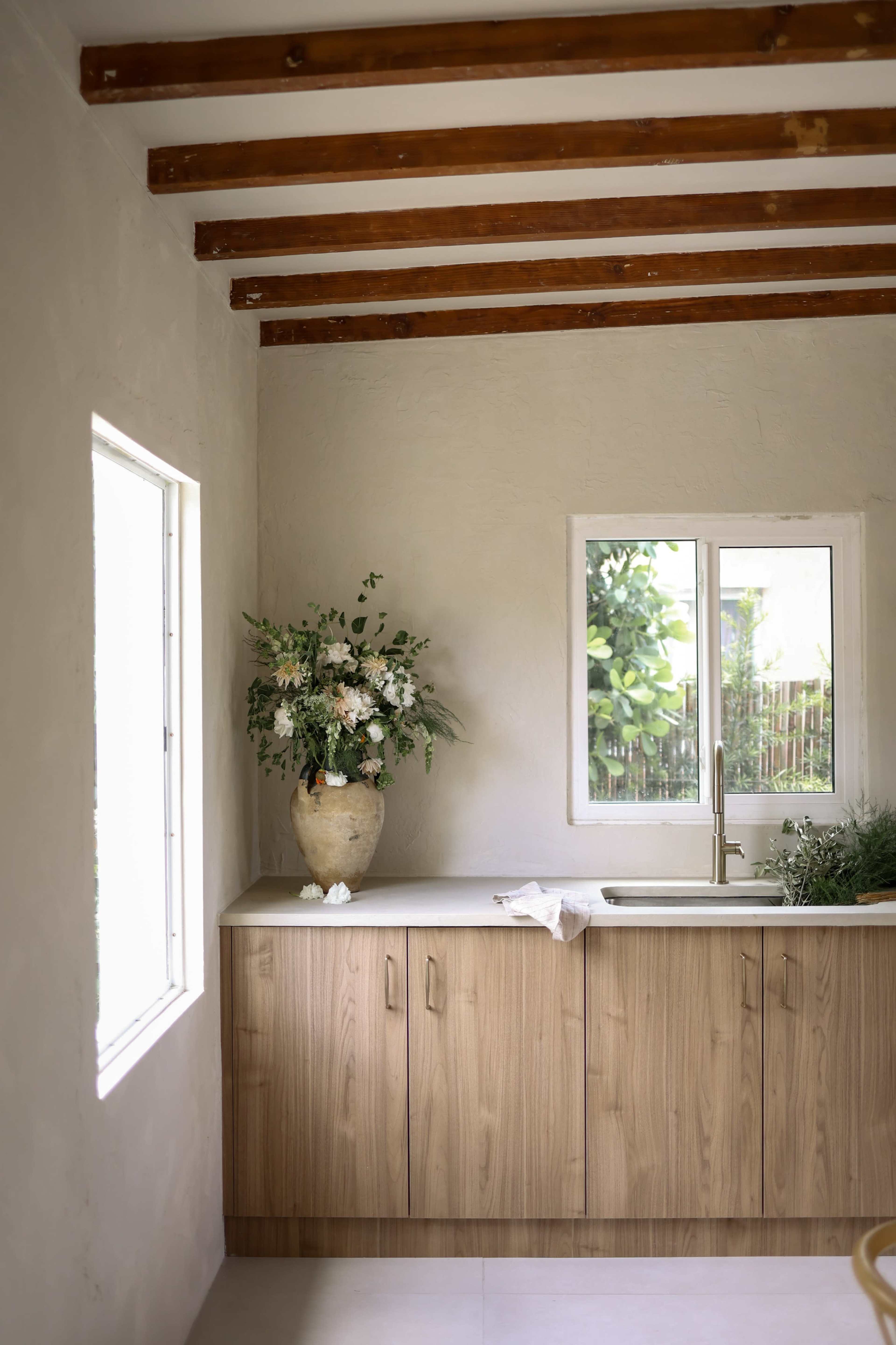 A modern kitchen features a wooden cabinetry, a sink, and a large vase with flowers placed on the countertop near a window.