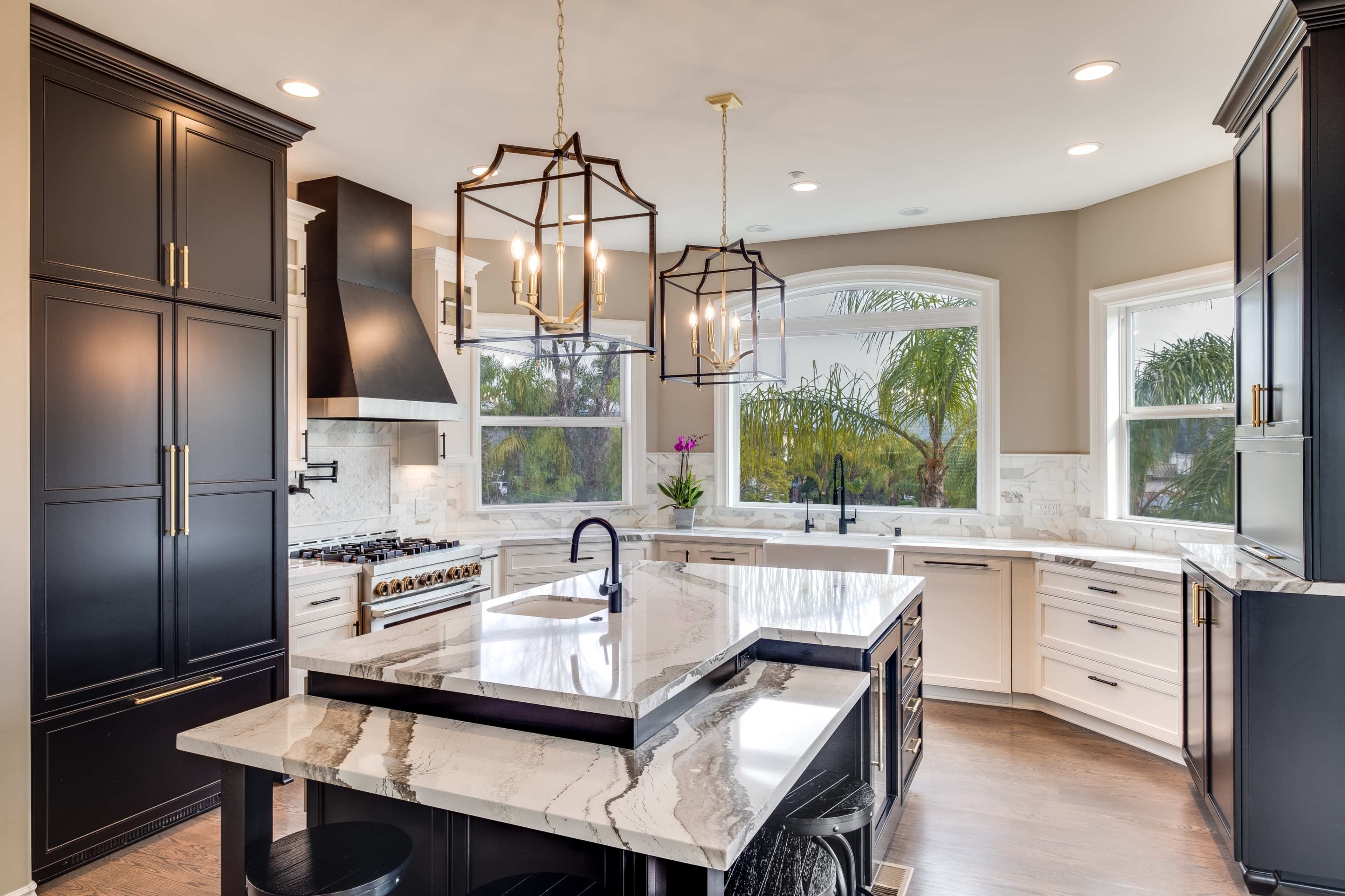The kitchen features a large marble island, black cabinetry, and a window overlooking palm trees.