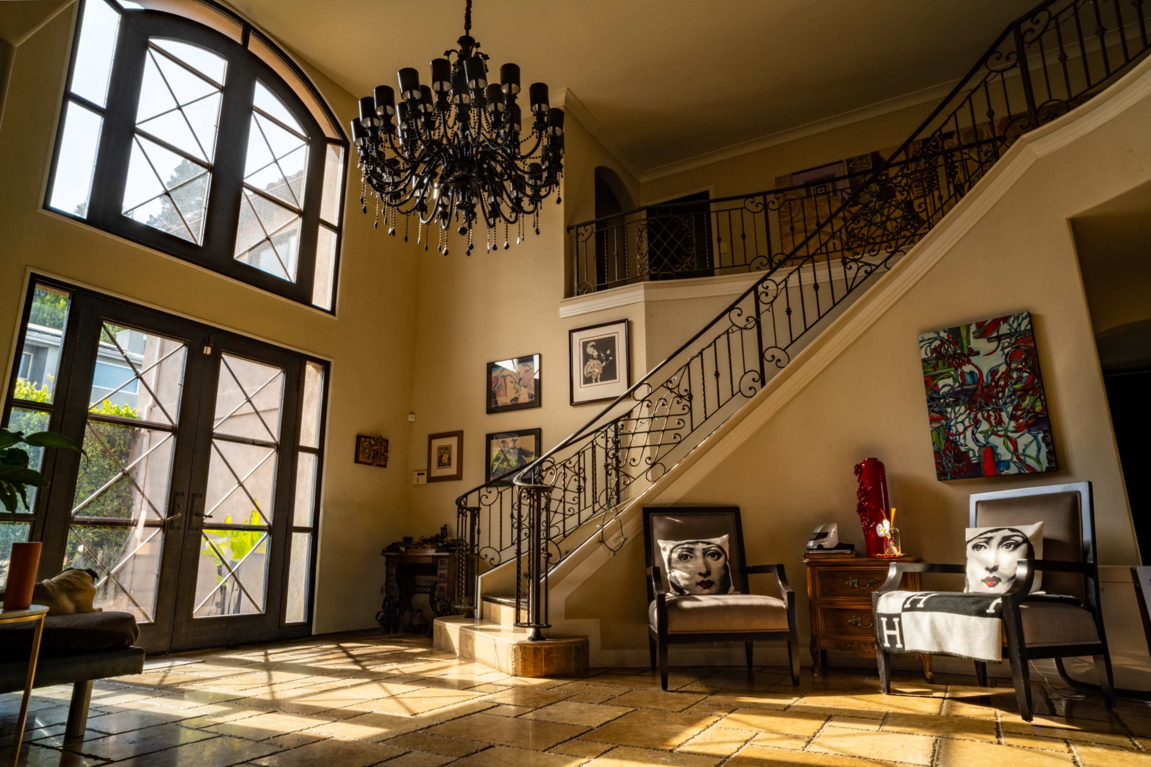 The image shows a well-lit entrance hall with a chandelier, a staircase, and stylish chairs featuring decorative face designs.