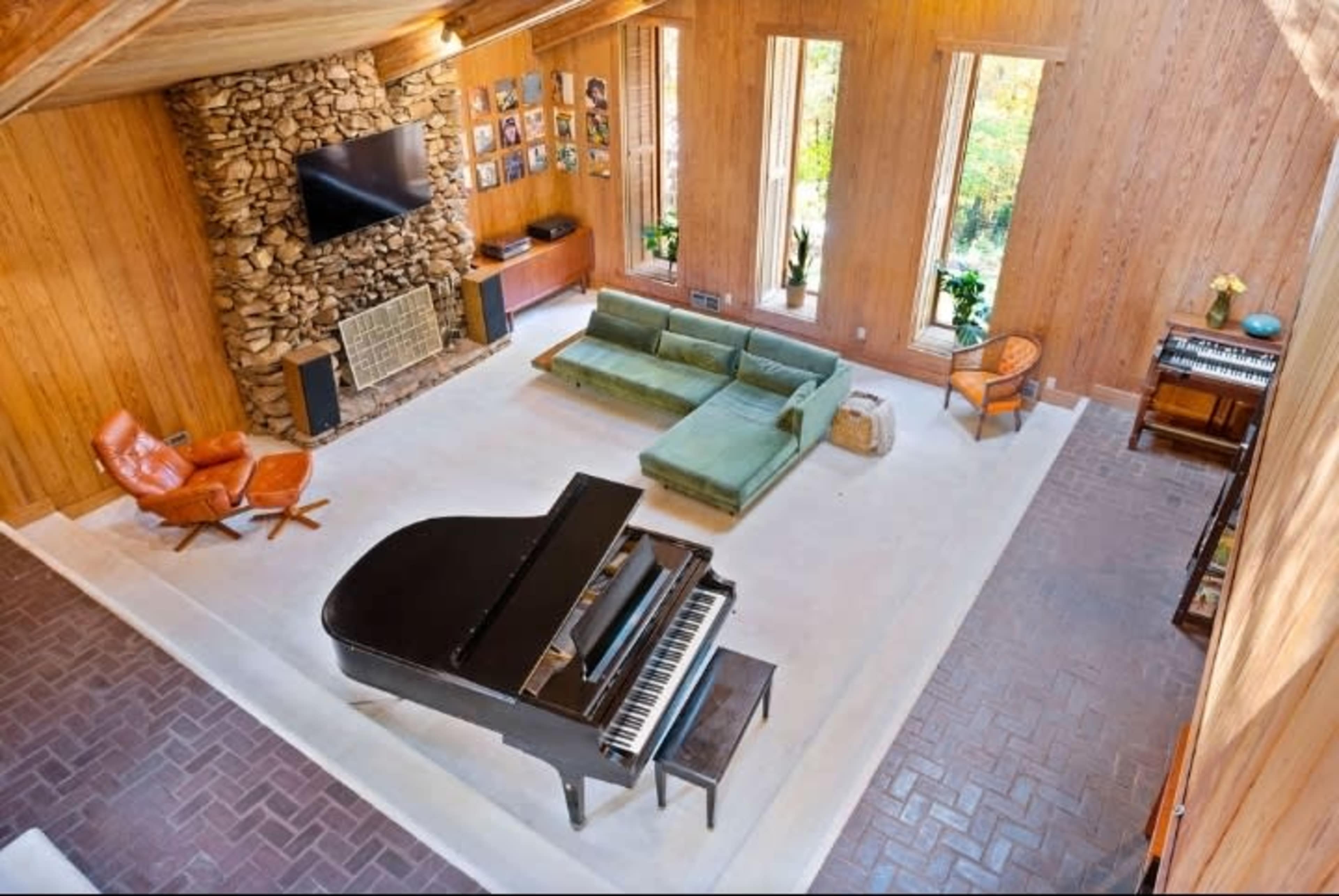 A spacious living room with a grand piano, green sofa, a stone fireplace, and large windows allowing natural light.