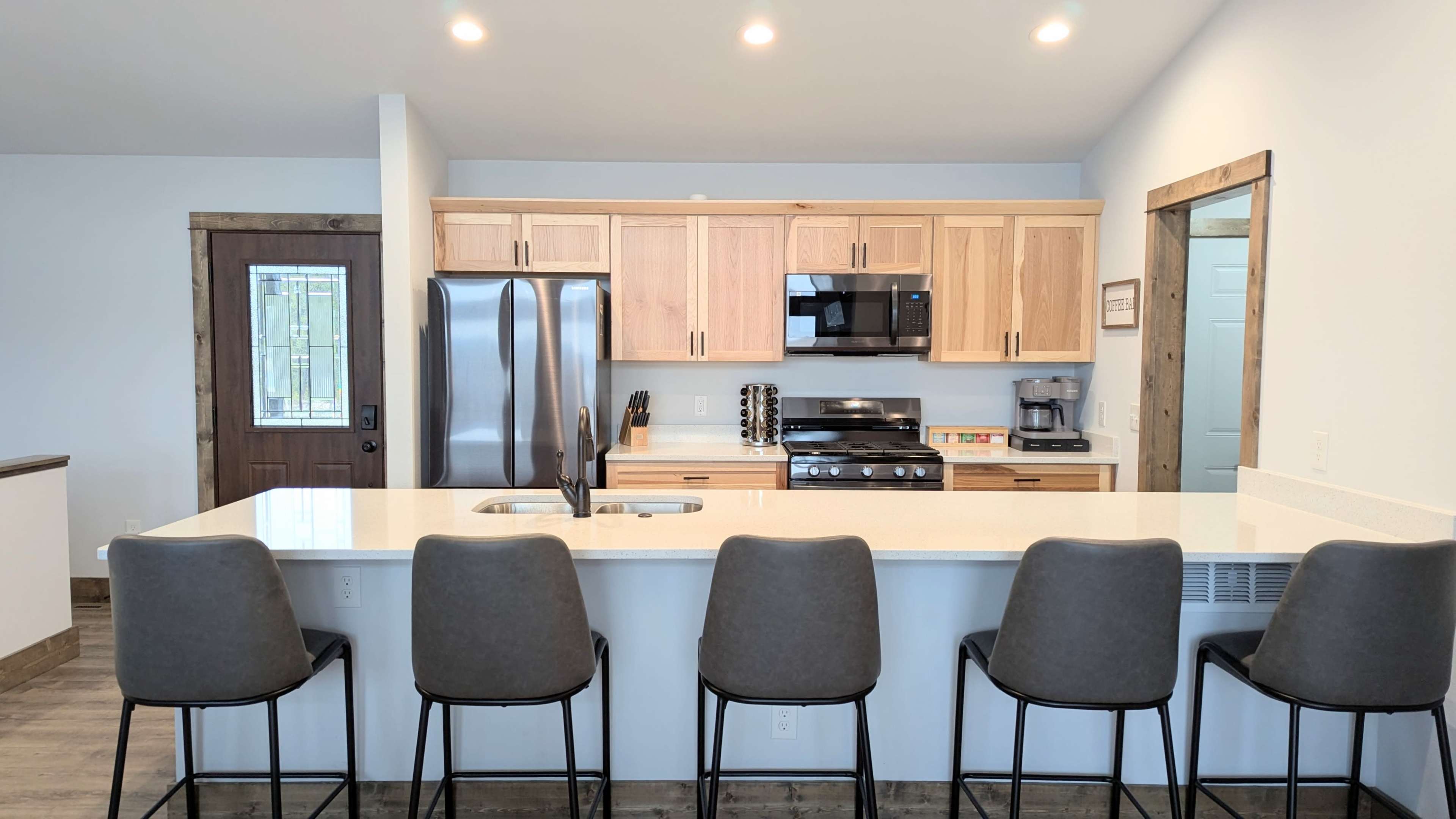 The image shows a modern kitchen with wooden cabinetry, stainless steel appliances, and a large island with seating for five.