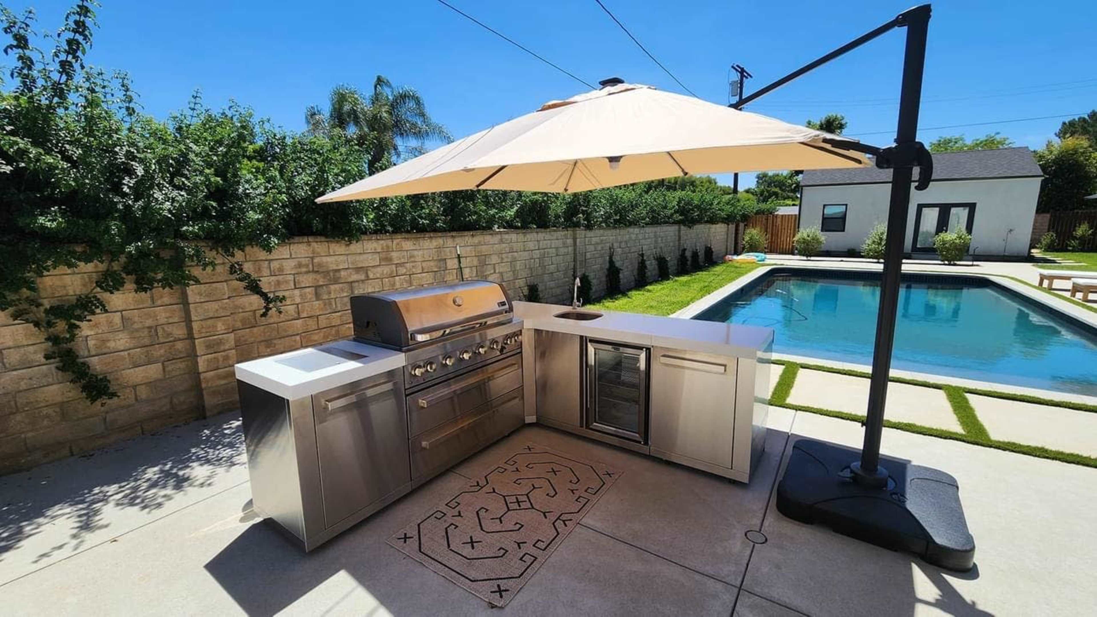 The image shows an outdoor kitchen with a grill, sink, and refrigerator next to a swimming pool, all shaded by a large umbrella.