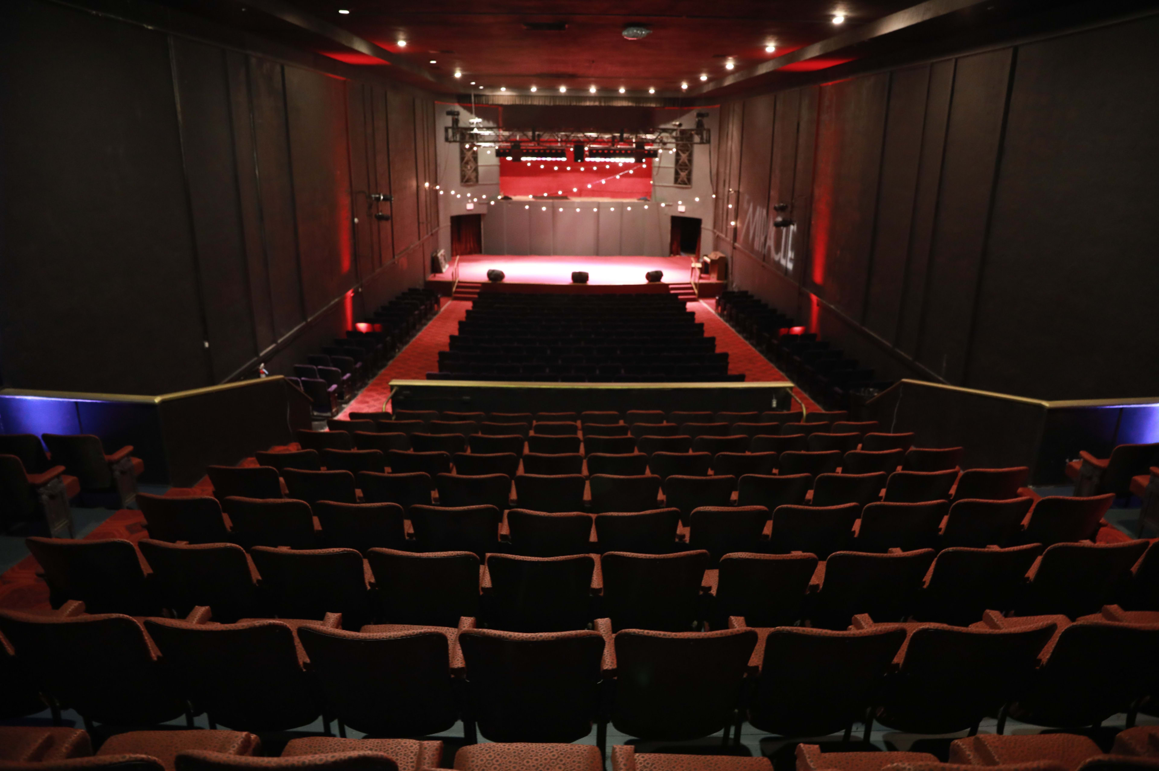 A theater auditorium with rows of empty red seats facing a well-lit stage at the front.