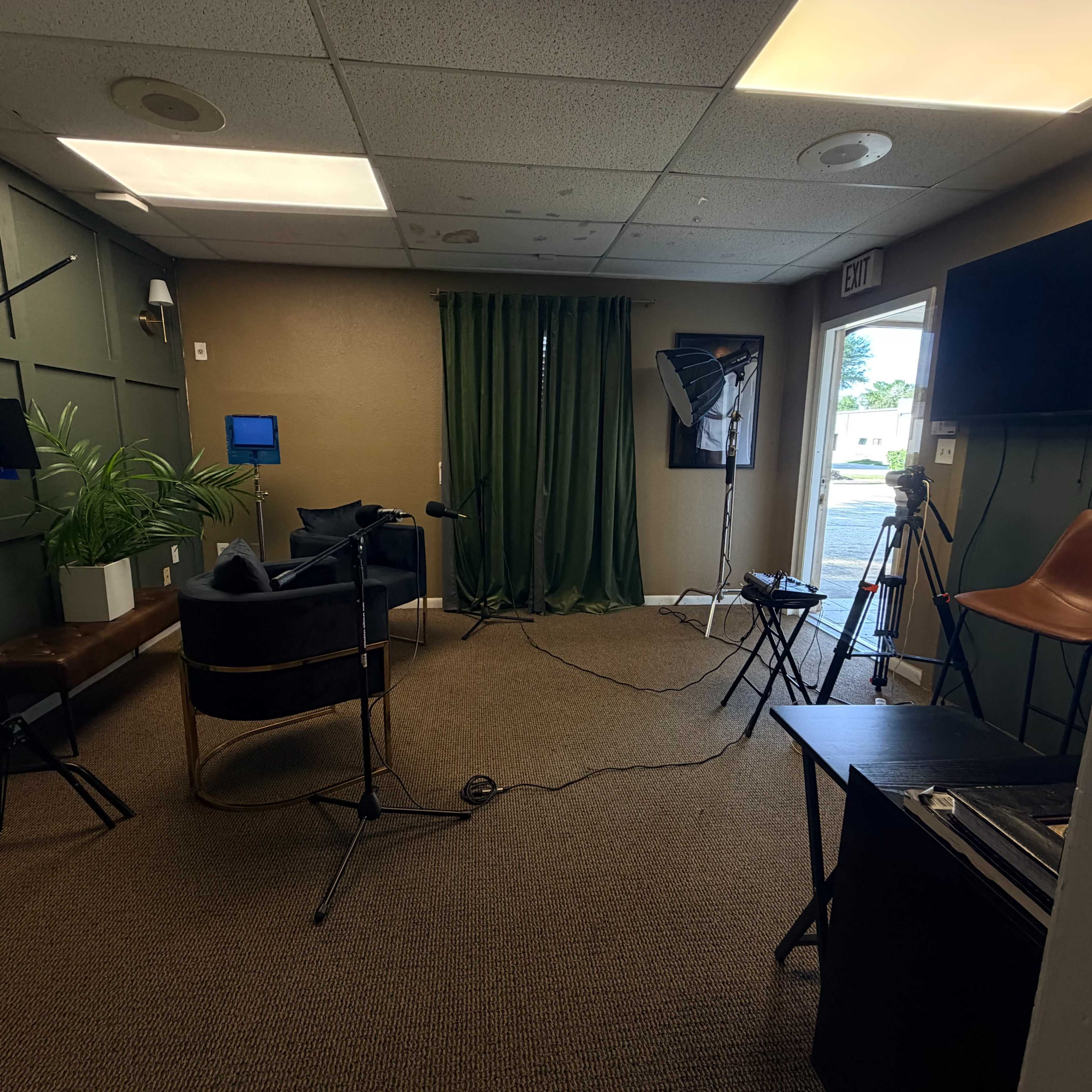 The image shows a small studio or interview space with green curtains, two chairs, a microphone setup, and cameras positioned near an exit door.