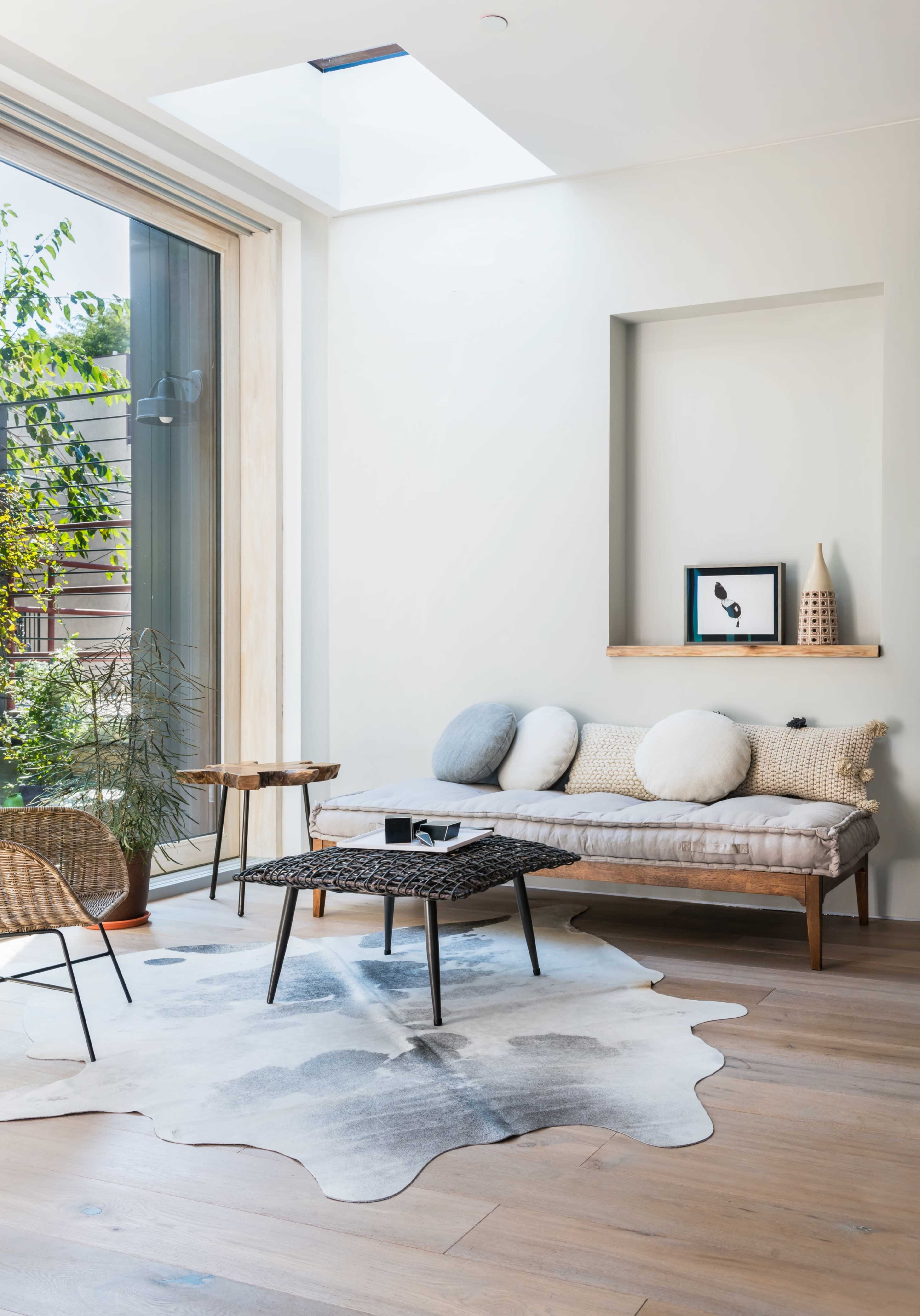 The image depicts a modern living room with a daybed, a low coffee table, a patterned rug, and large windows allowing natural light to illuminate the space.