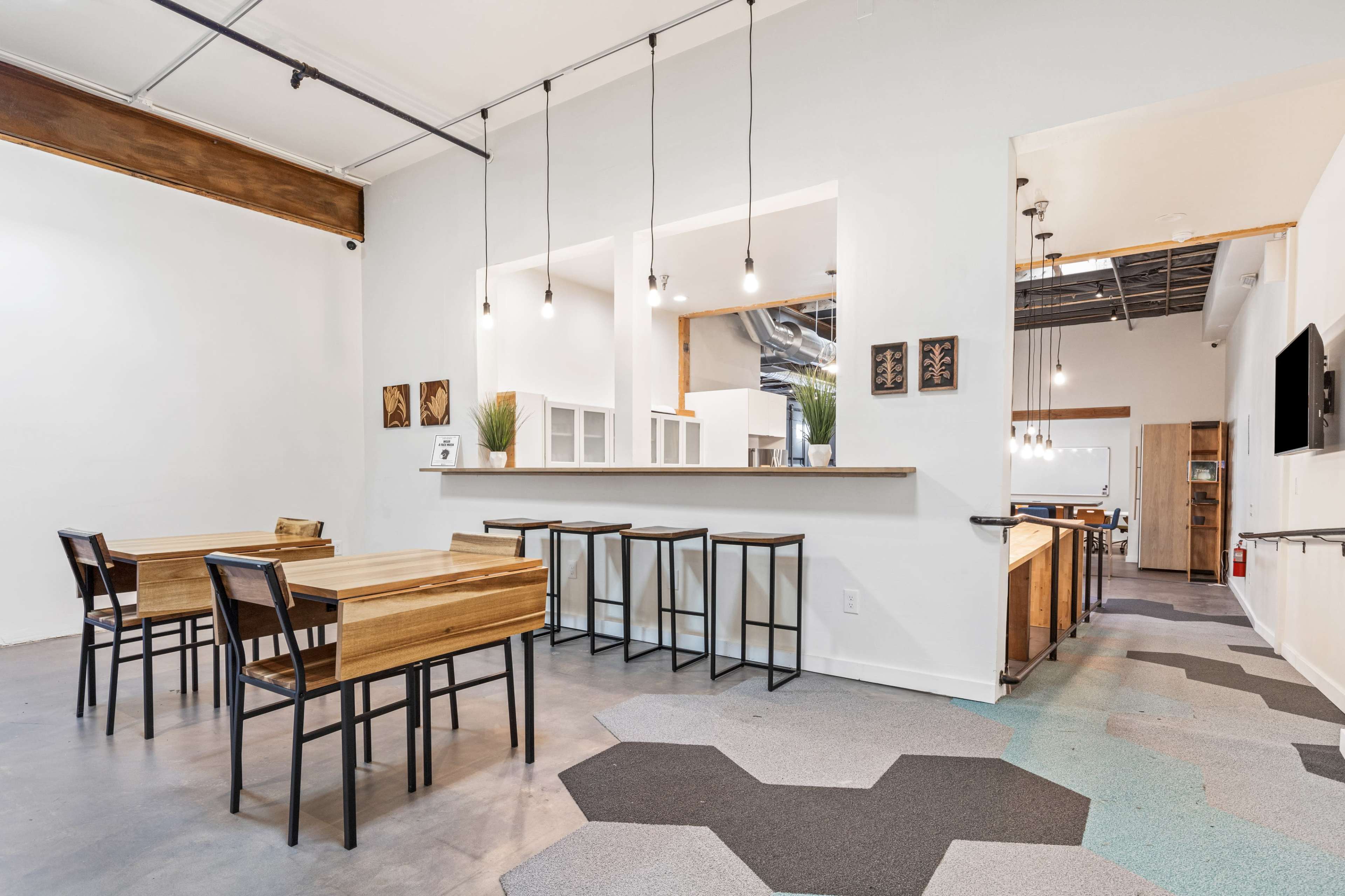 The image shows a minimalist café interior with wooden furniture, including tables and stools, set against a neutral-colored wall and a patterned floor.