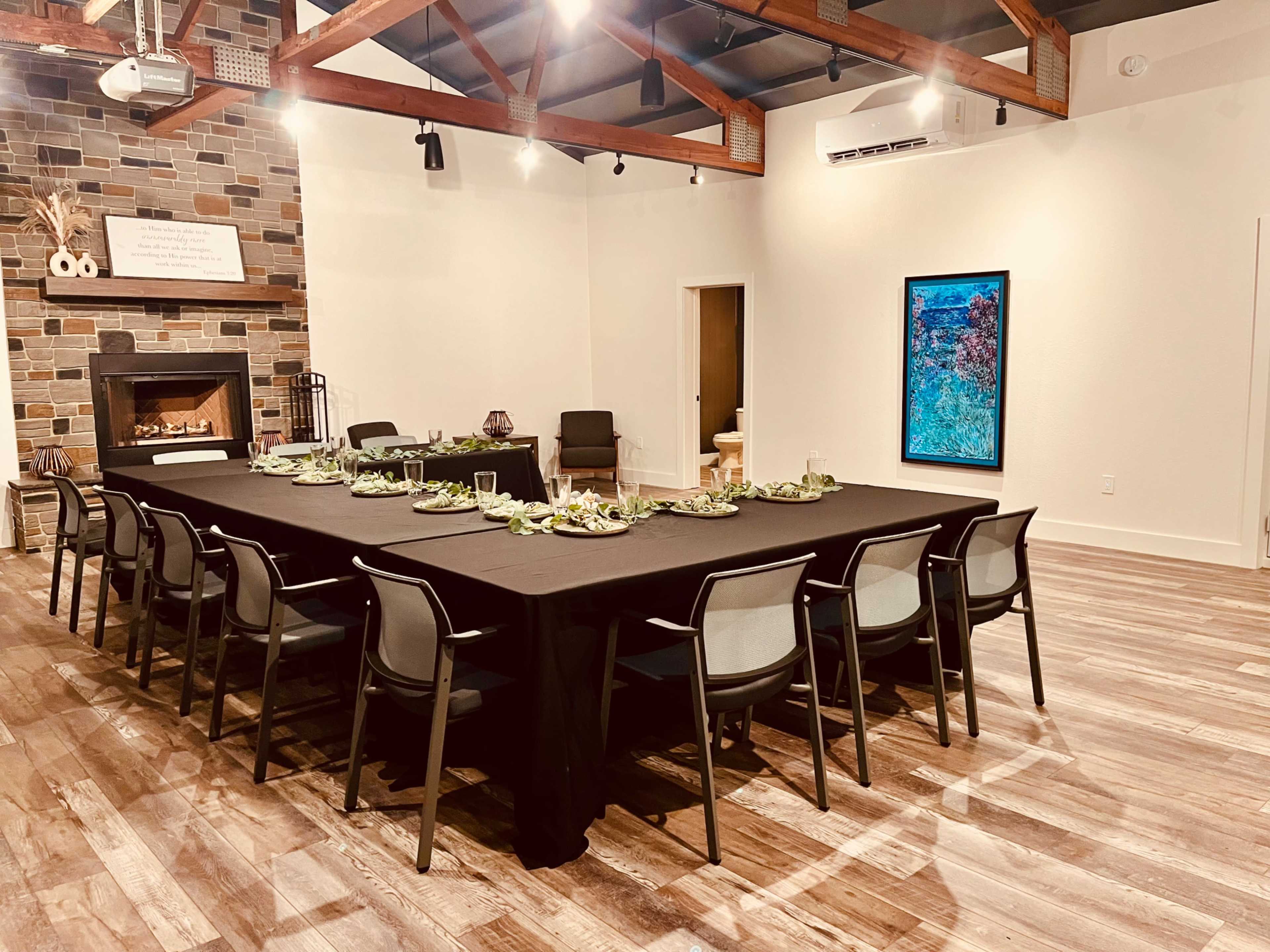 A long table is set with plates and decorations in a well-lit room featuring a stone fireplace and exposed wooden beams.