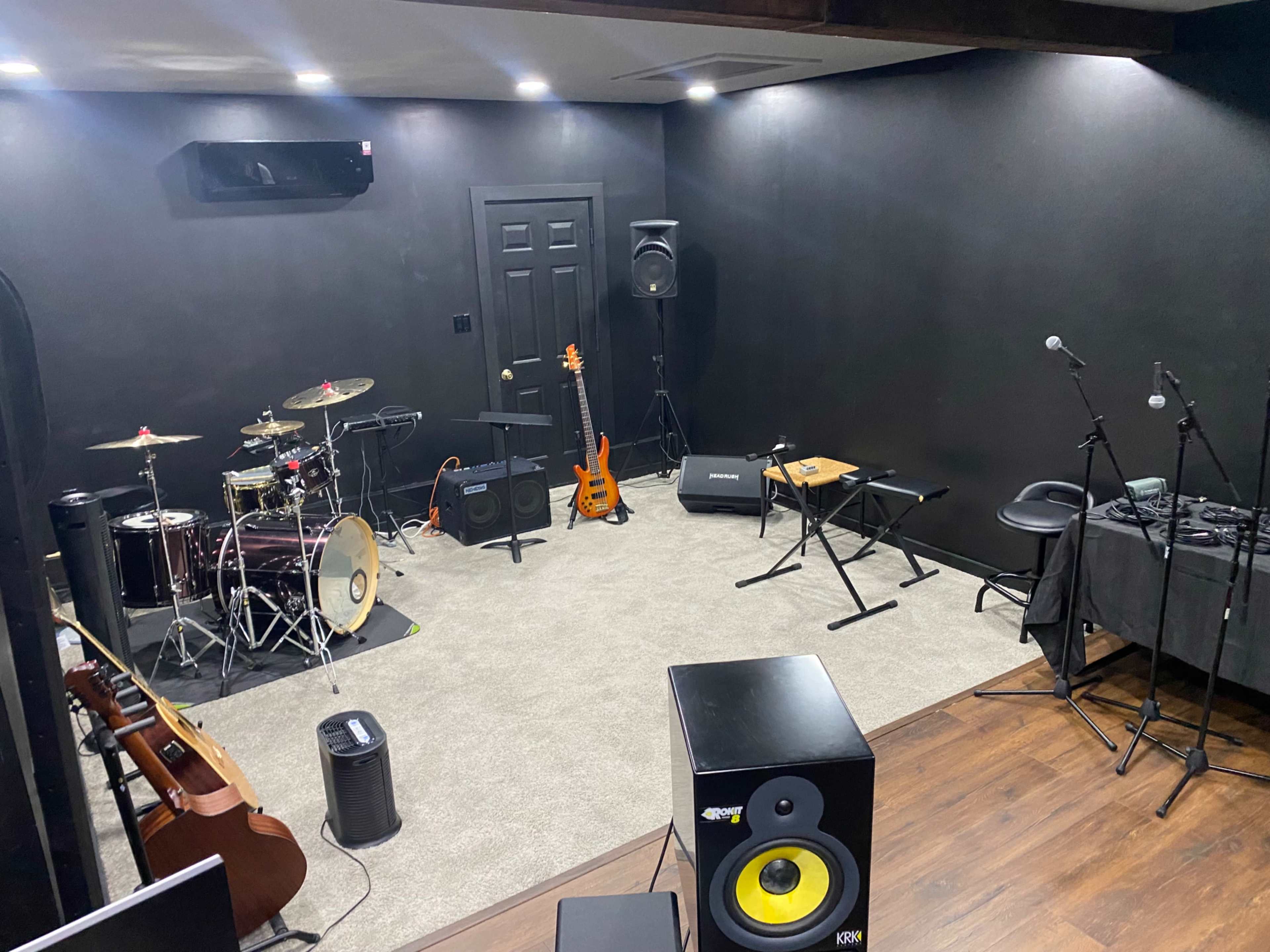 The image shows a music rehearsal studio equipped with various instruments, including a drum set, electric guitars, and a sound mixing board, along with microphones and amplifiers in a soundproofed room.