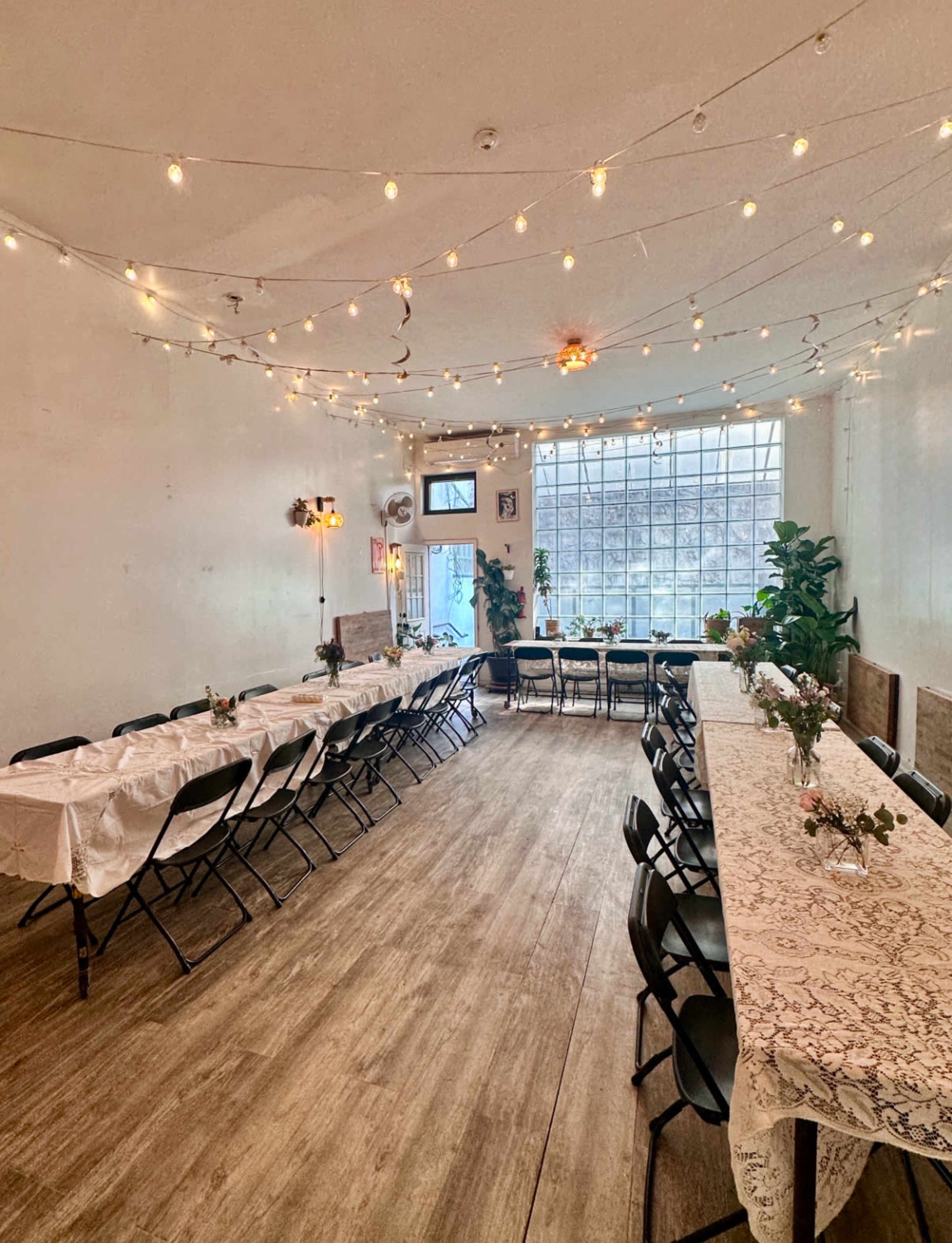 The image shows a well-lit event space with long tables set with lace tablecloths and small floral centerpieces, surrounded by folding chairs and string lights overhead.