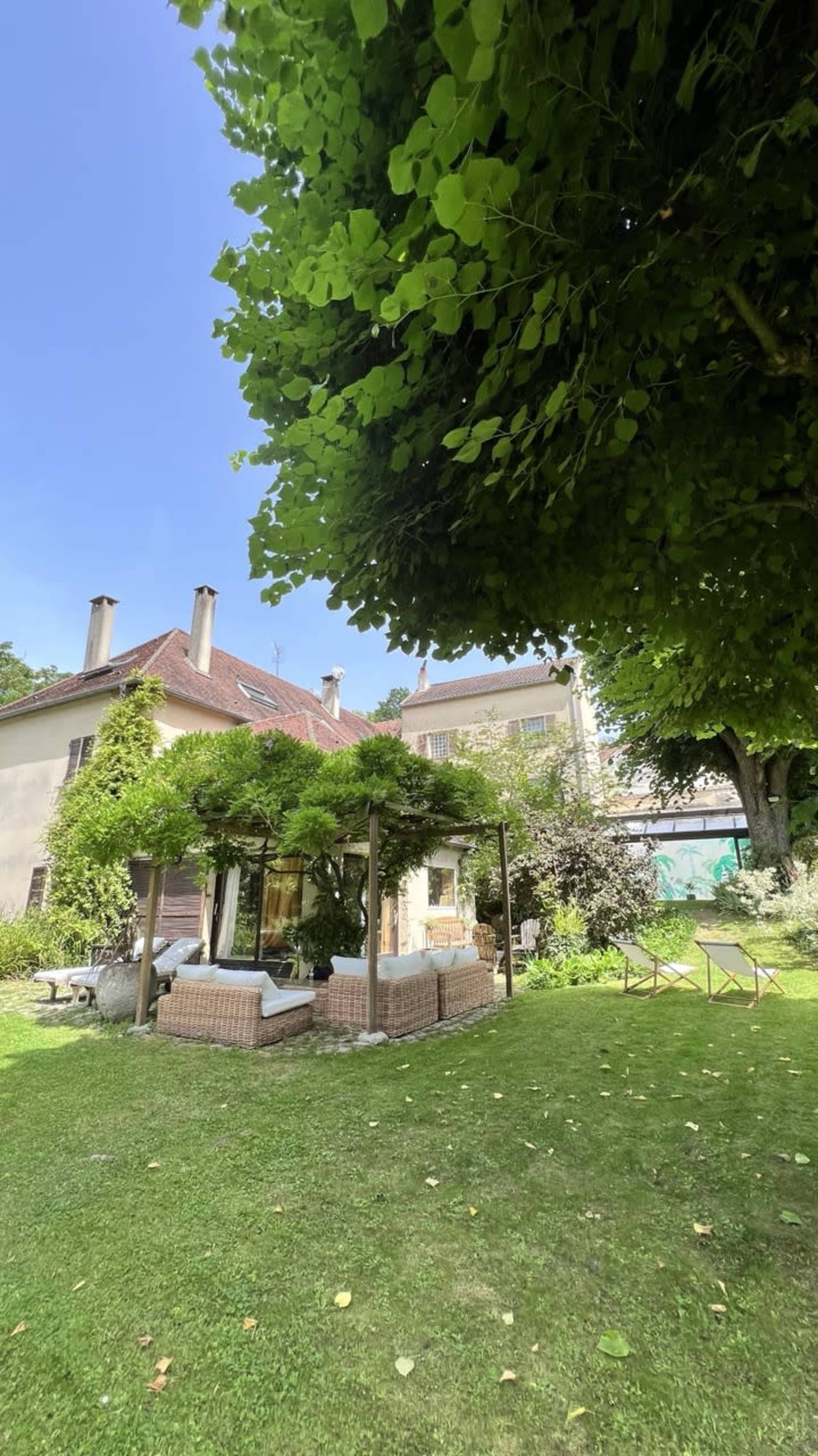 A spacious garden features a seating area under shaded trees, with a view of a modern house in the background.