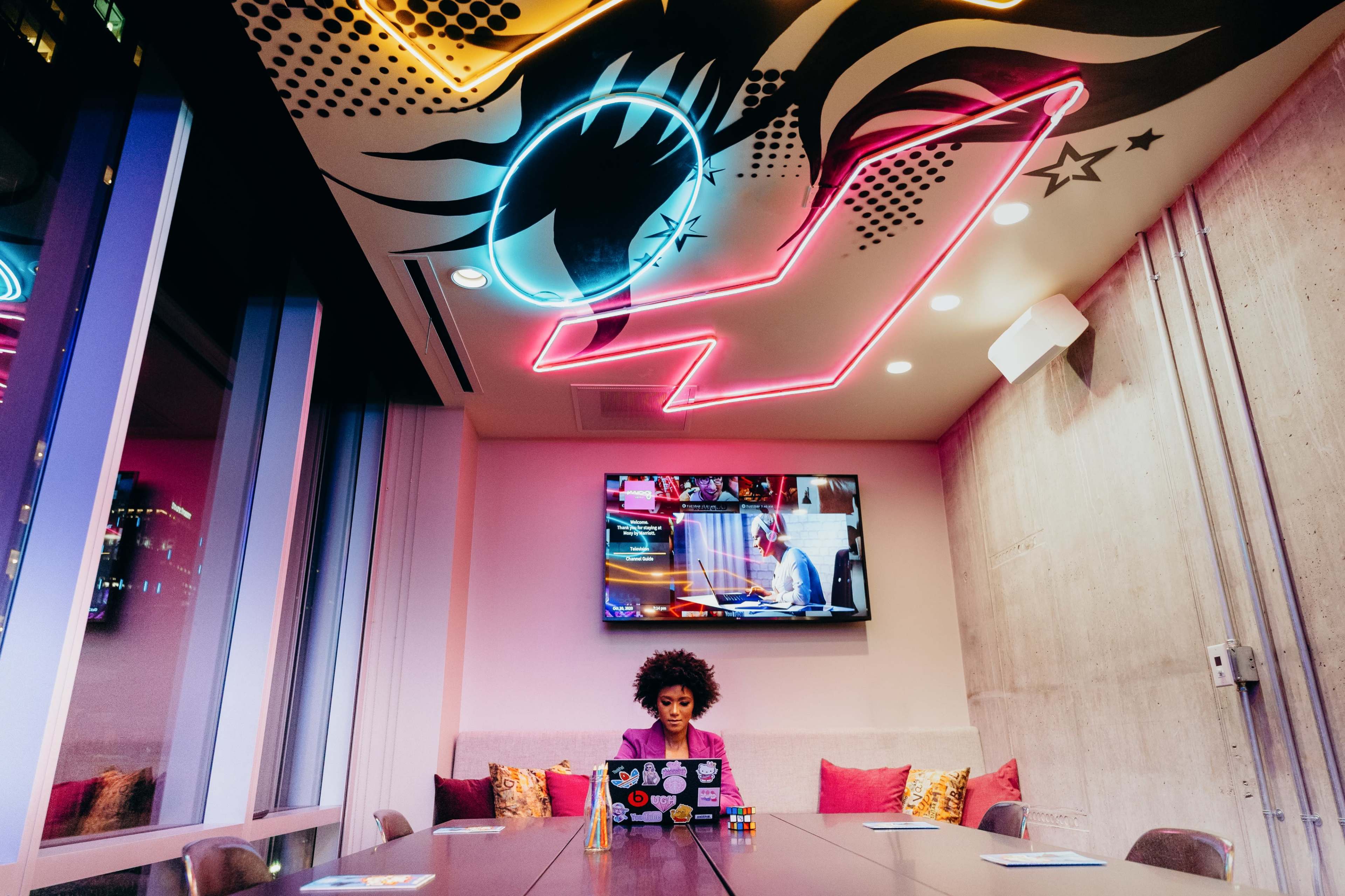A woman sits at a long table in a brightly lit conference room with colorful graffiti on the ceiling and a television displaying a video in the background.