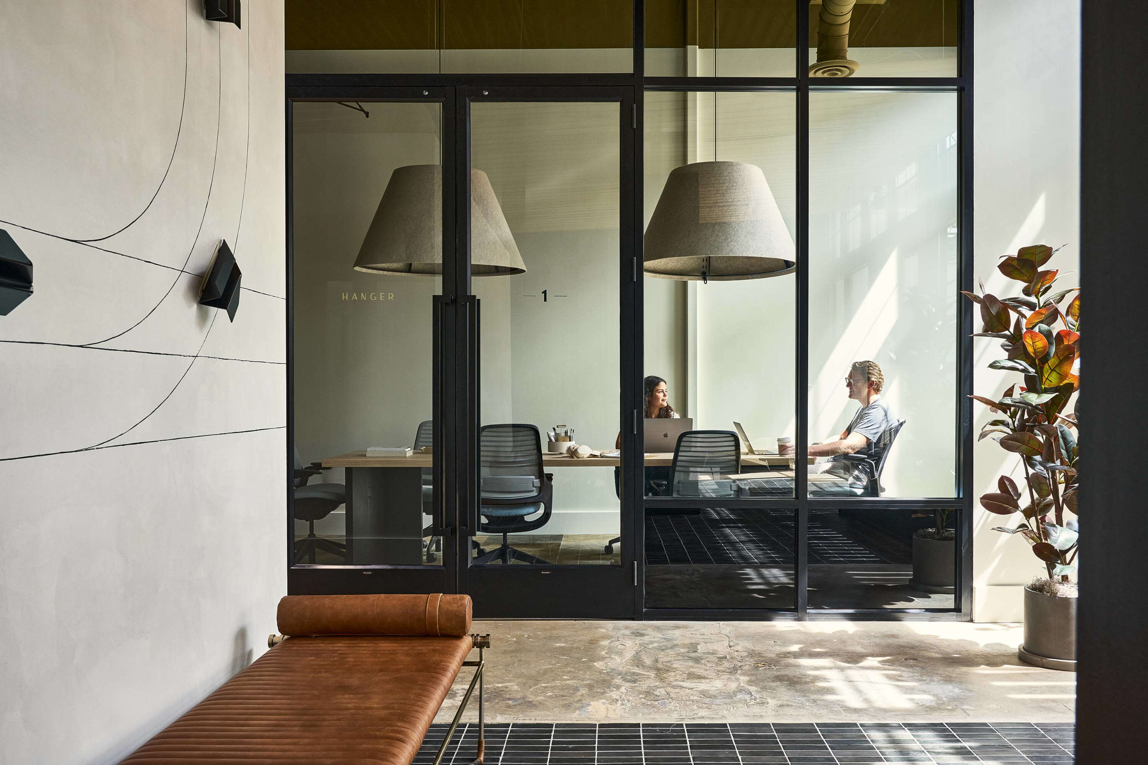 Two people are seated at a conference table in a glass-walled meeting room, with large pendant lamps overhead and a potted plant visible in the foreground.
