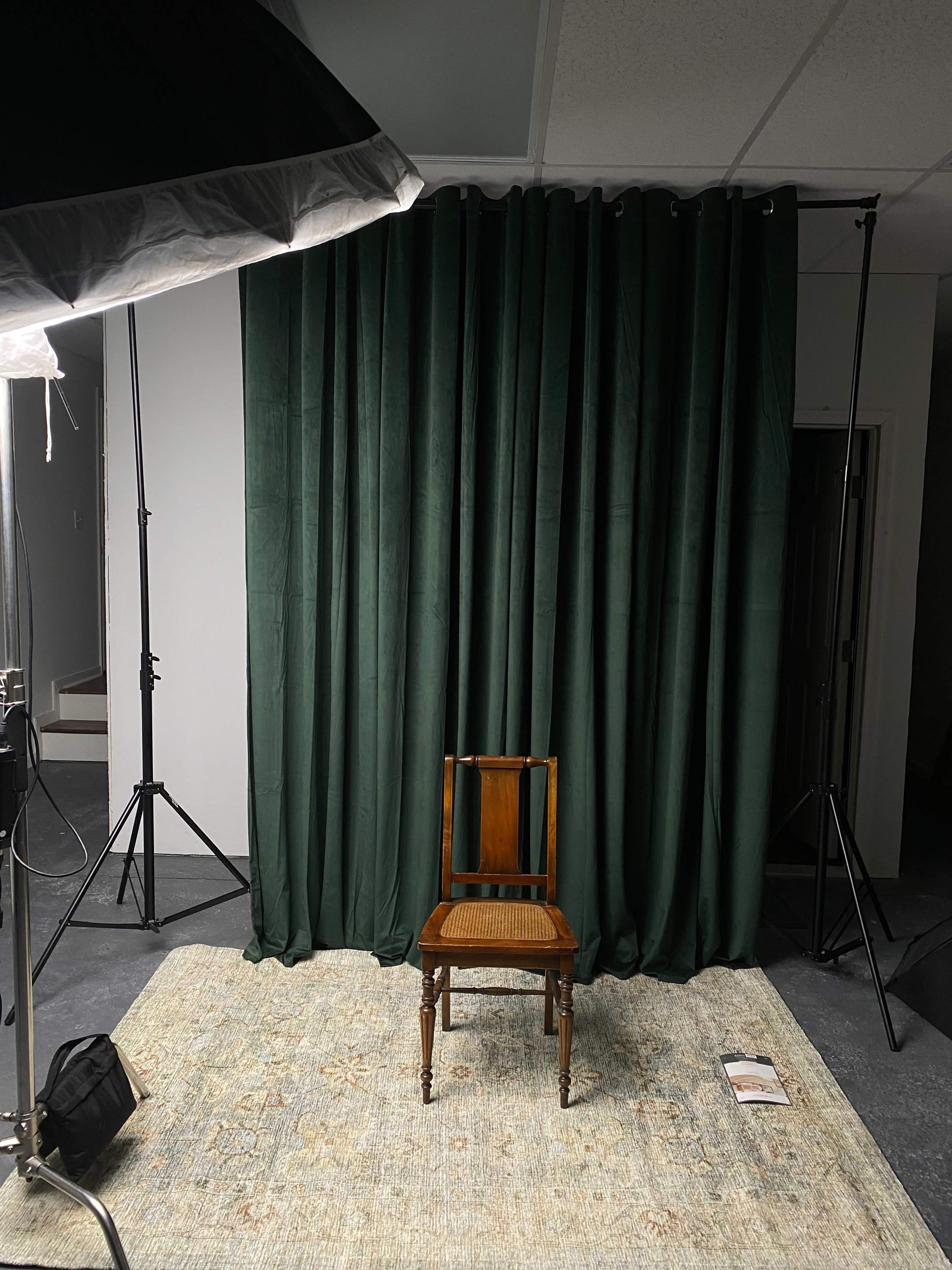 A single wooden chair is centered in front of a dark green curtain, surrounded by studio lighting equipment on a patterned rug.