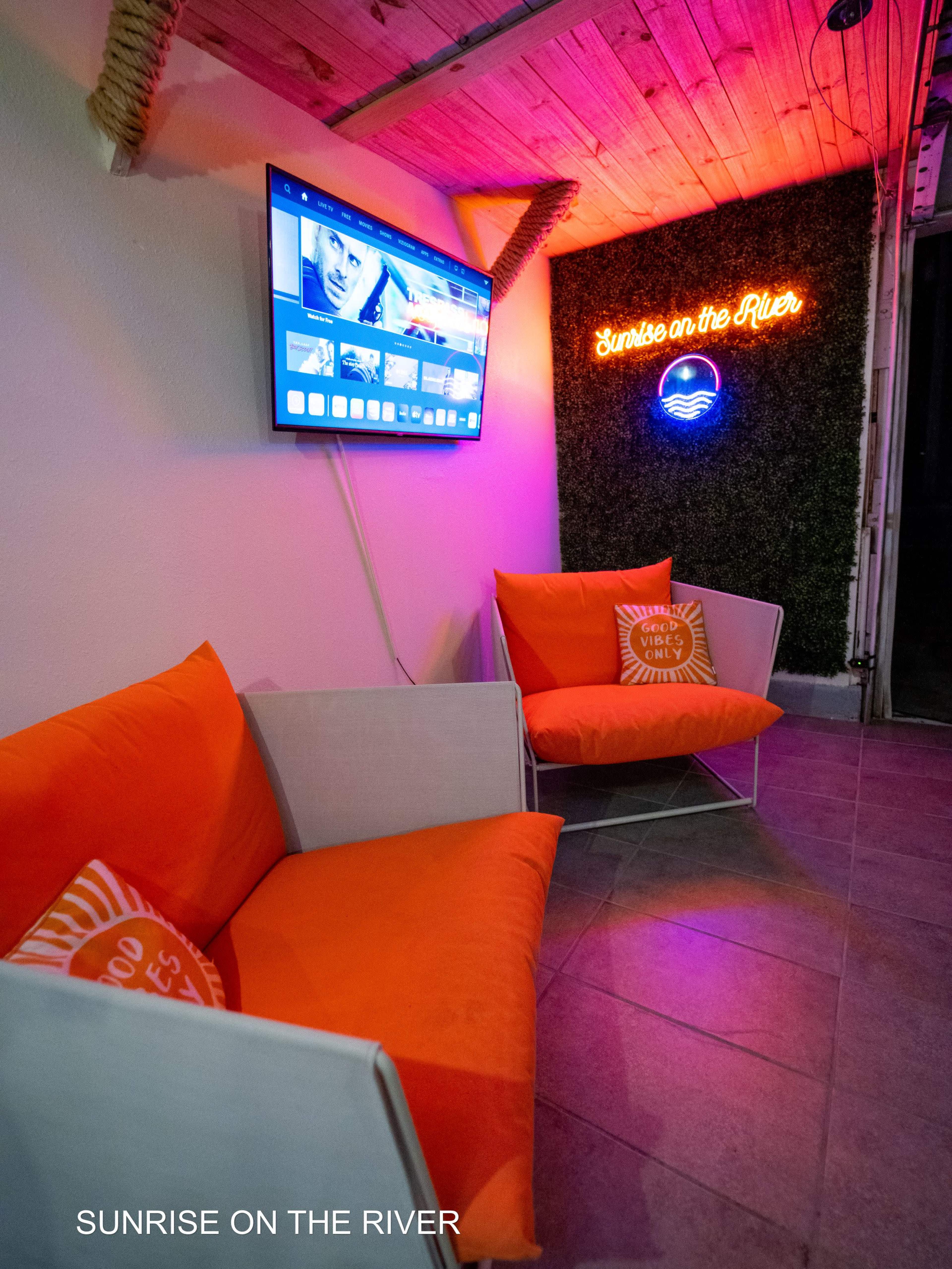 A cozy seating area with two orange chairs, a television mounted on the wall, and a glowing neon sign that reads "Sunrise on the River."