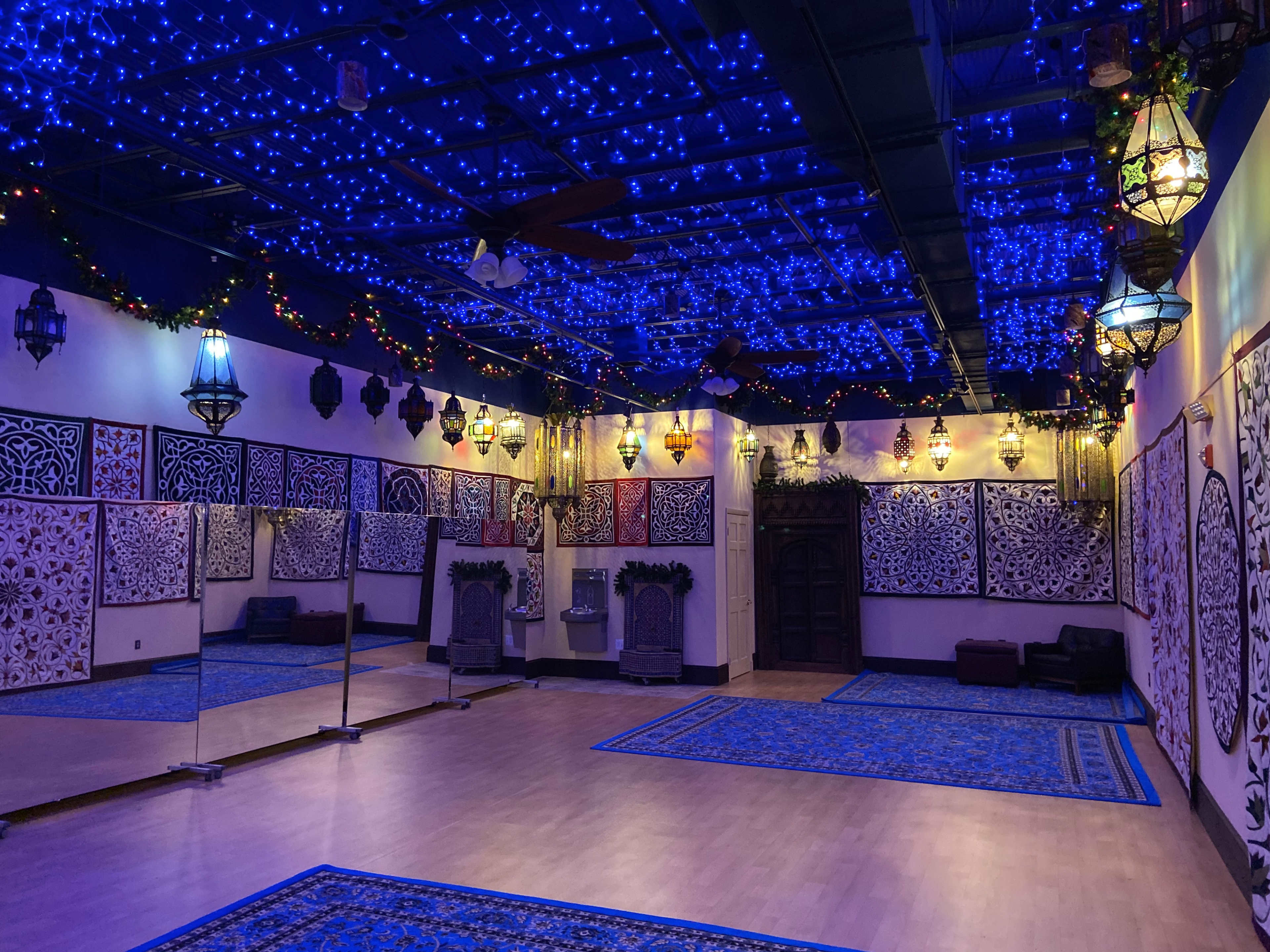 The image shows an interior space decorated with blue string lights, ornate lanterns, and patterned wall hangings, featuring wooden benches and large mirrors.