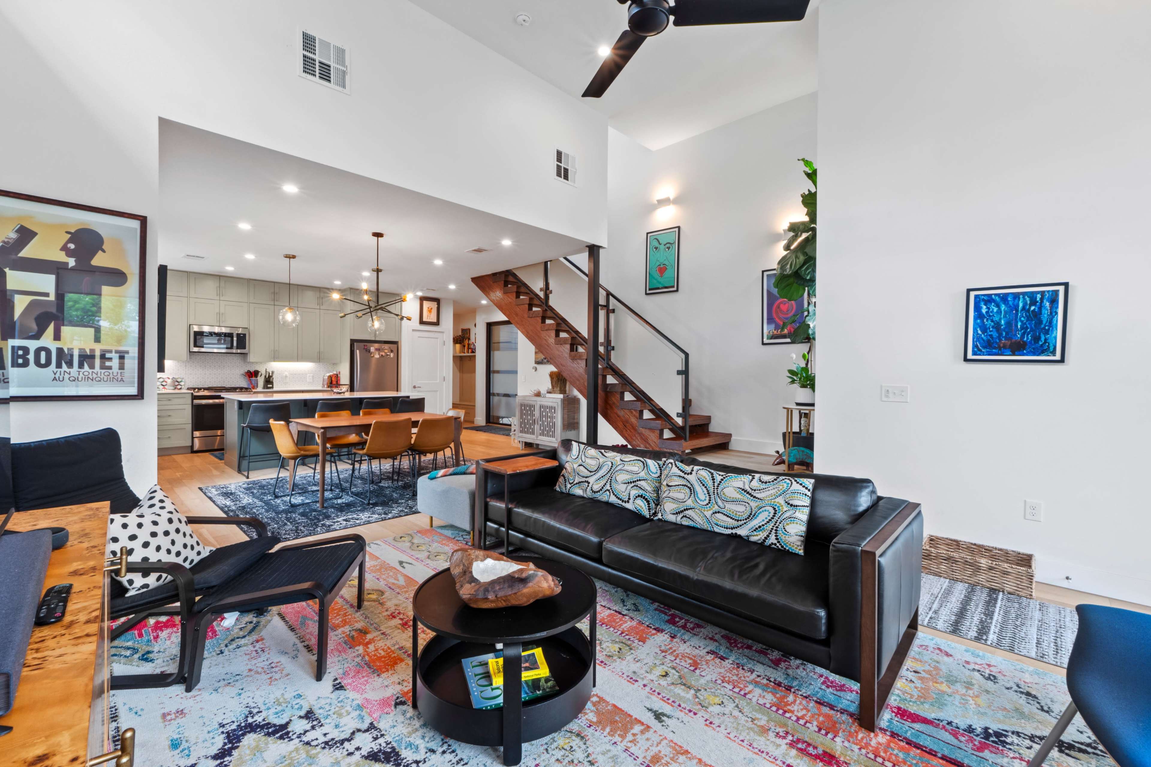 The image shows a modern living room with a staircase, a leather couch, and a dining area adjacent to a kitchen, illuminated by pendant lights.